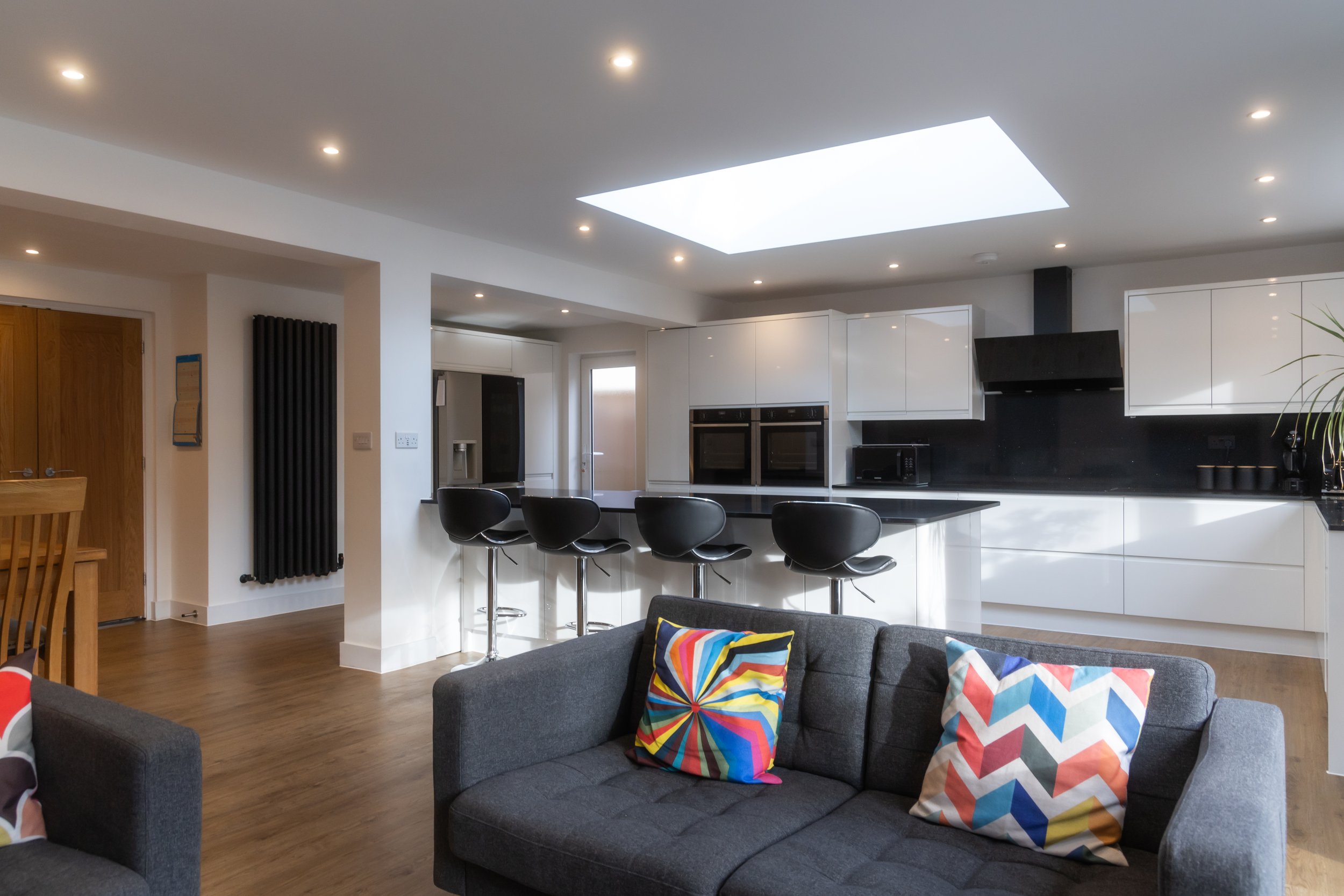 Modern kitchen and living room with white cabinets, black countertop, kitchen island with black barstools, gray sofa with colorful patterned pillows, and skylight in ceiling.