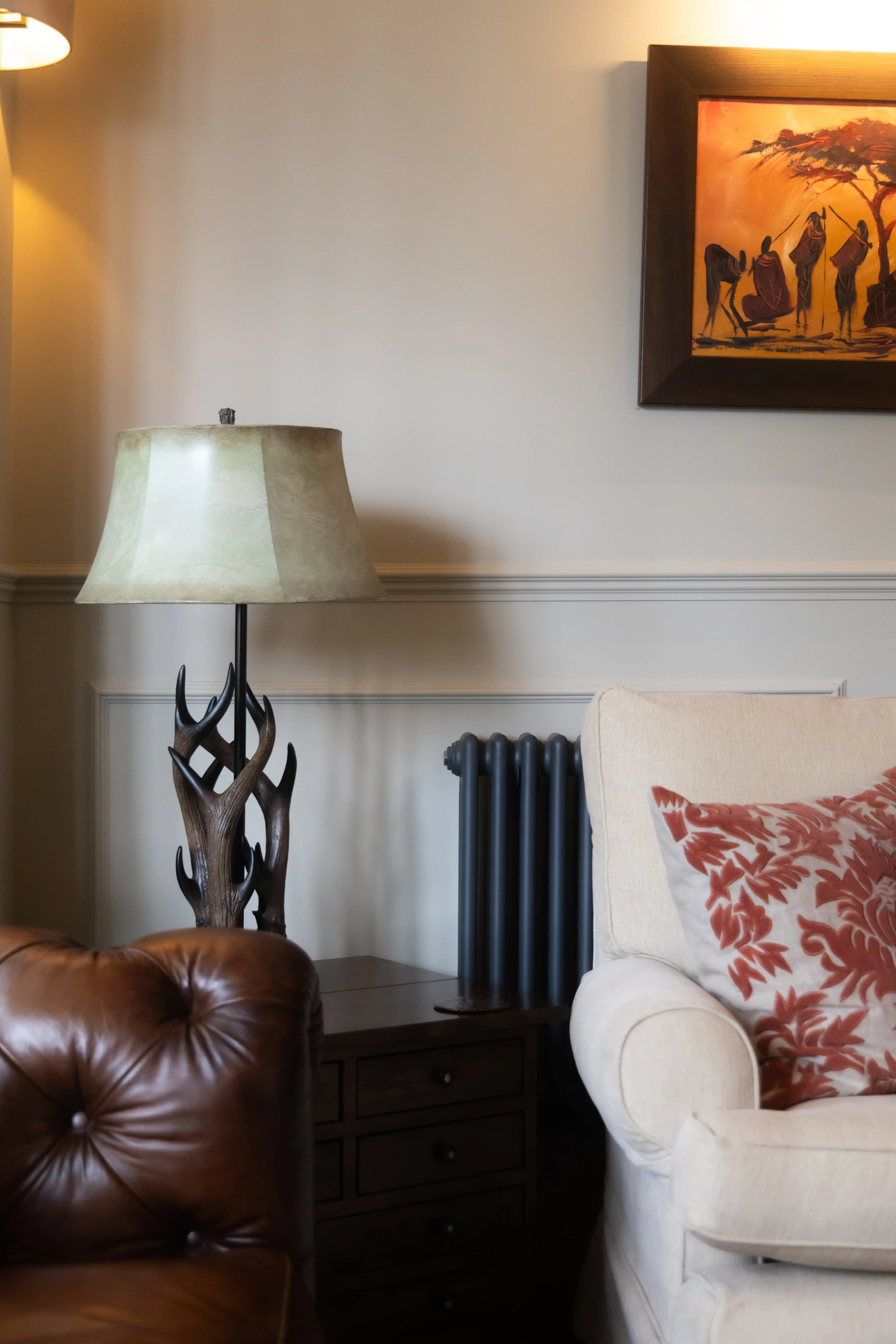 Living room corner with a lamp on a side table, a leather armchair, a platform bed with a white and orange pillow, a wall with a painting of African figures, and a radiator.