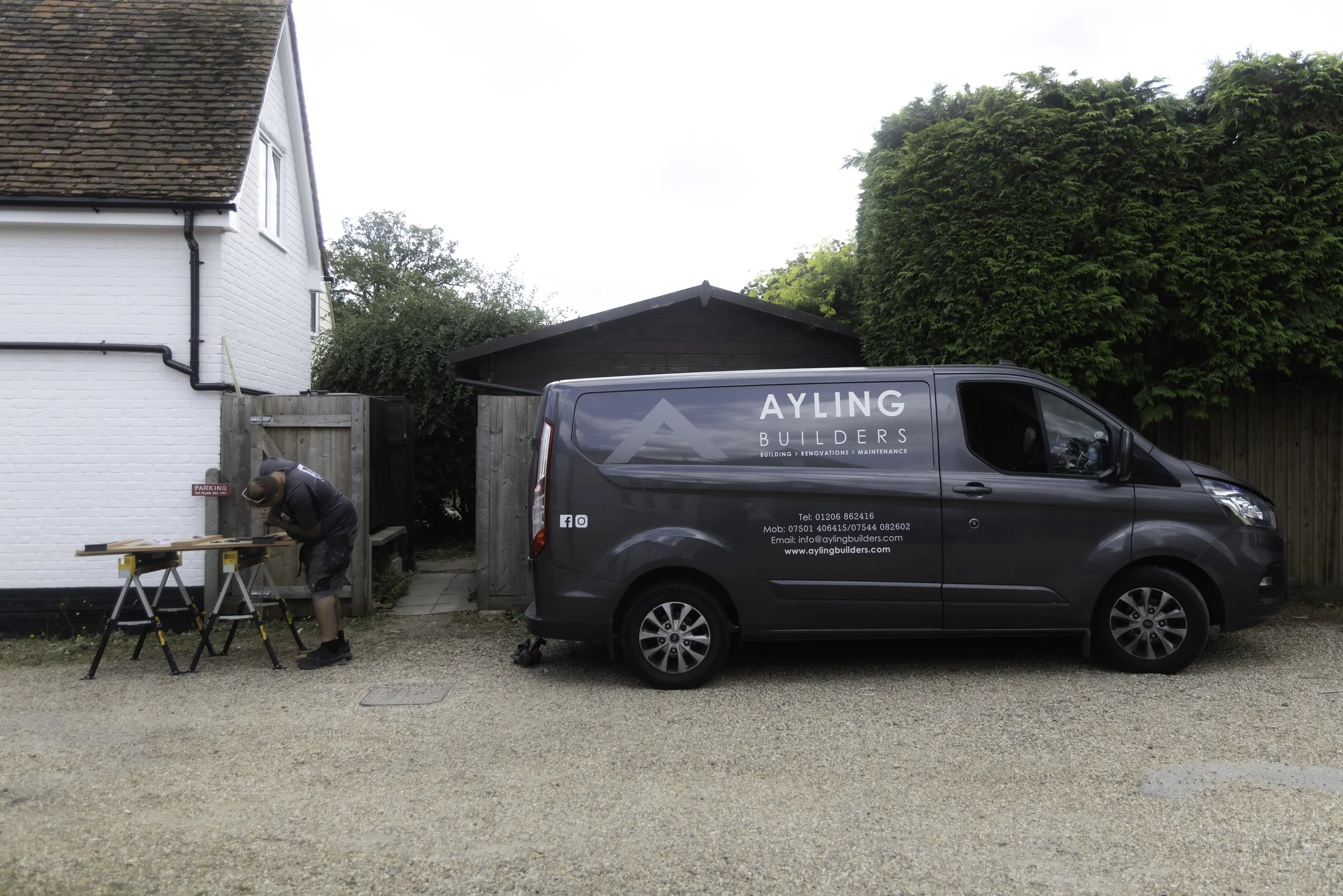 A man working at a construction site outside a residential house with a gray van parked nearby. The van has the logo and contact information of Aylings Builders.