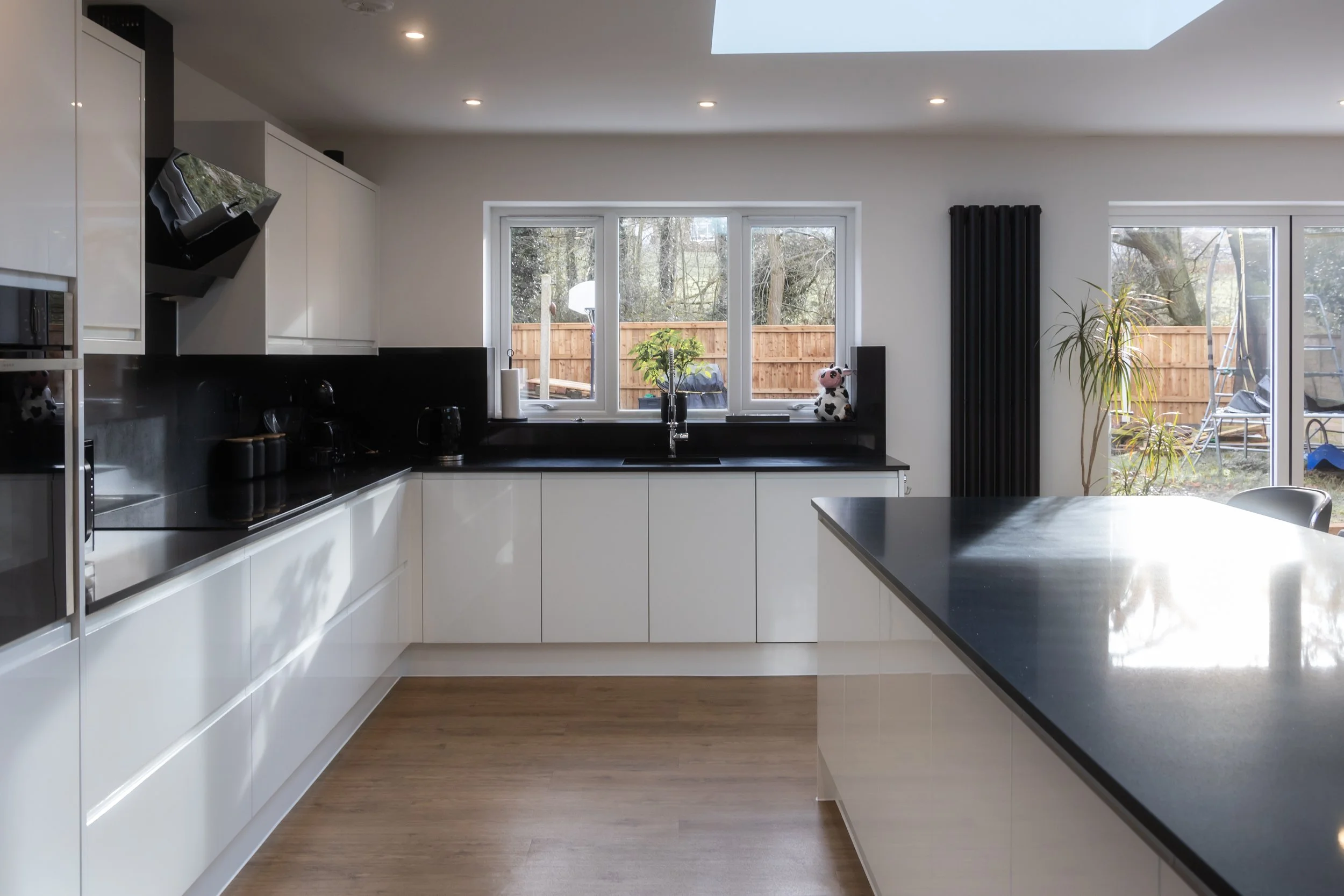 Modern kitchen with white cabinets, black countertops, large windows, and a view of the backyard with outdoor furniture and a wooden fence.