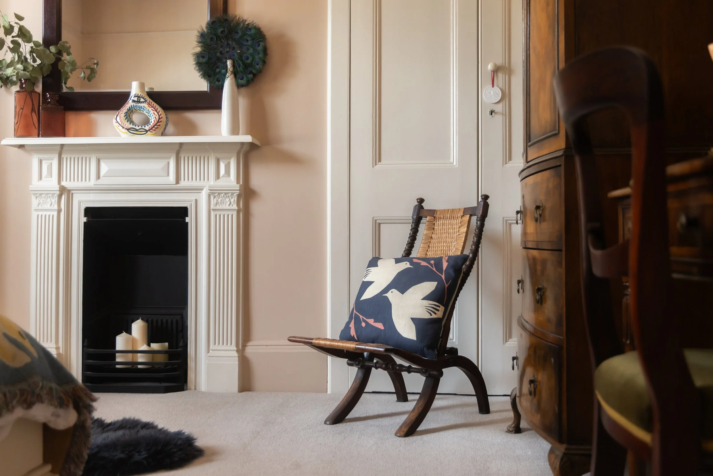 A cozy living room corner featuring a wooden chair with a cushion decorated with a bird design, a white fireplace mantle with candles, a peacock feather wreath on the wall, a large mirror, and a wooden cabinet.