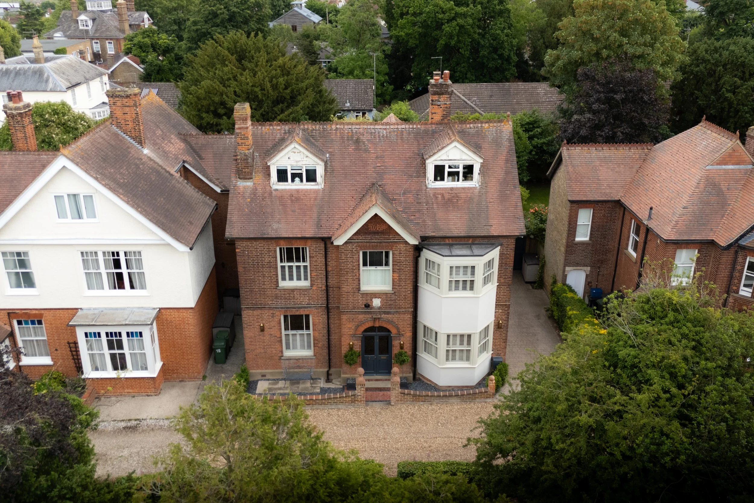 Aerial view of a row of older brick houses with chimneys, pitched roofs, and bay windows, surrounded by mature trees and greenery in a suburban neighborhood.