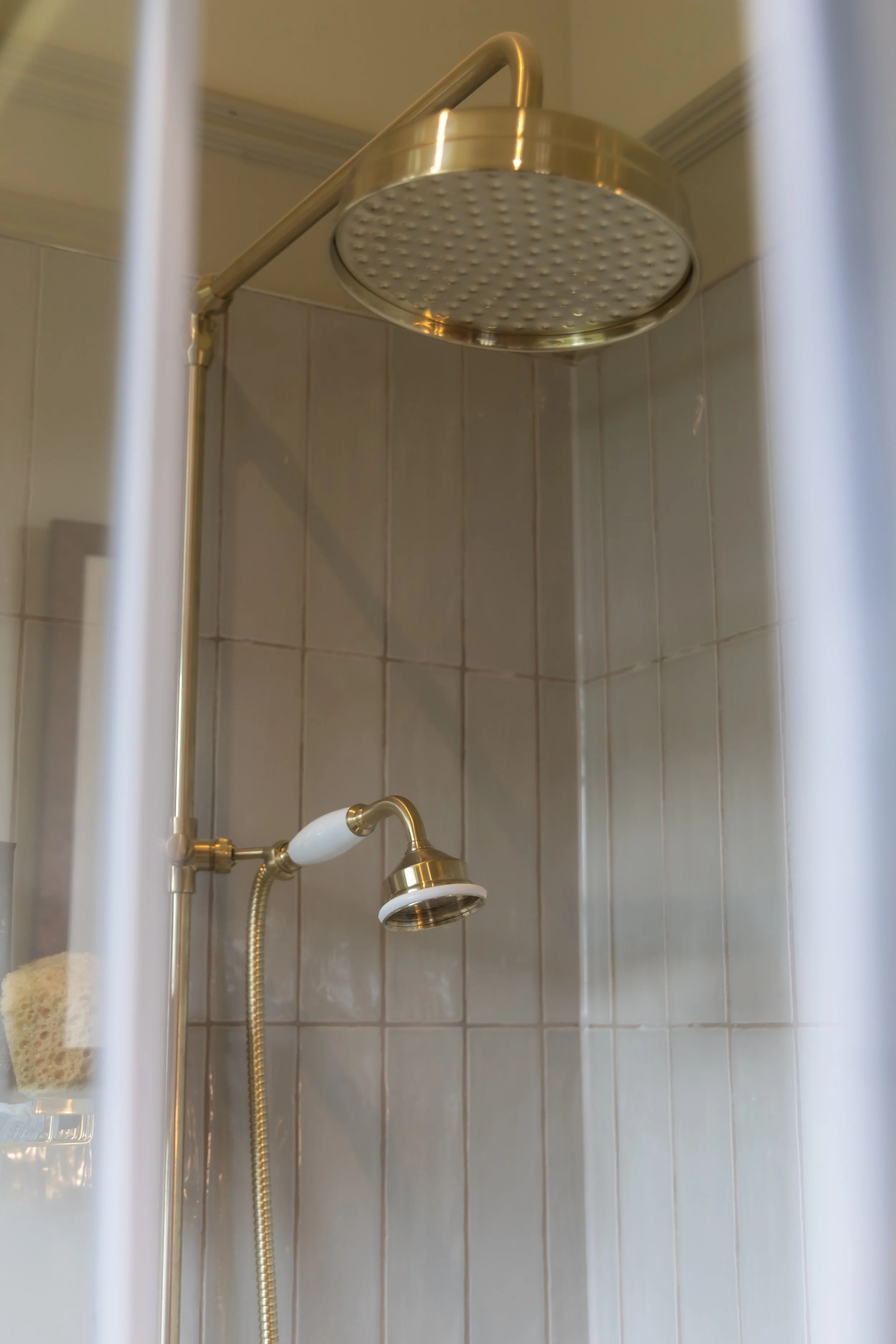 Shower with brass fixtures, beige tiled walls, foam sponge and loofah hanging, and a soap dispenser on a shelf.
