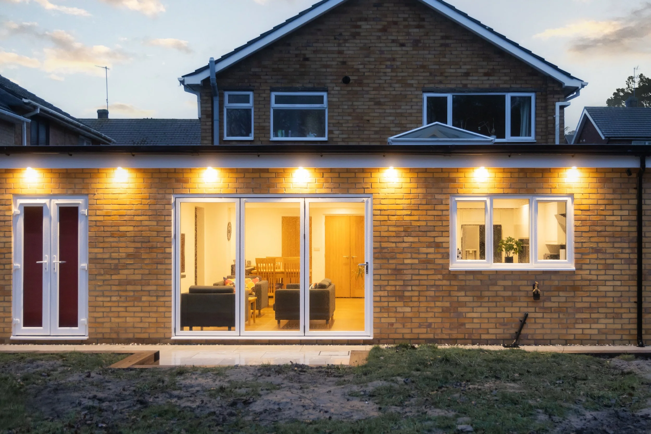 The image shows the back of a brick house during dusk, with the interior lights on. The house has large sliding glass doors opening into a living room with chairs and a wooden dining table. The backyard has a patchy lawn and a concrete patio.