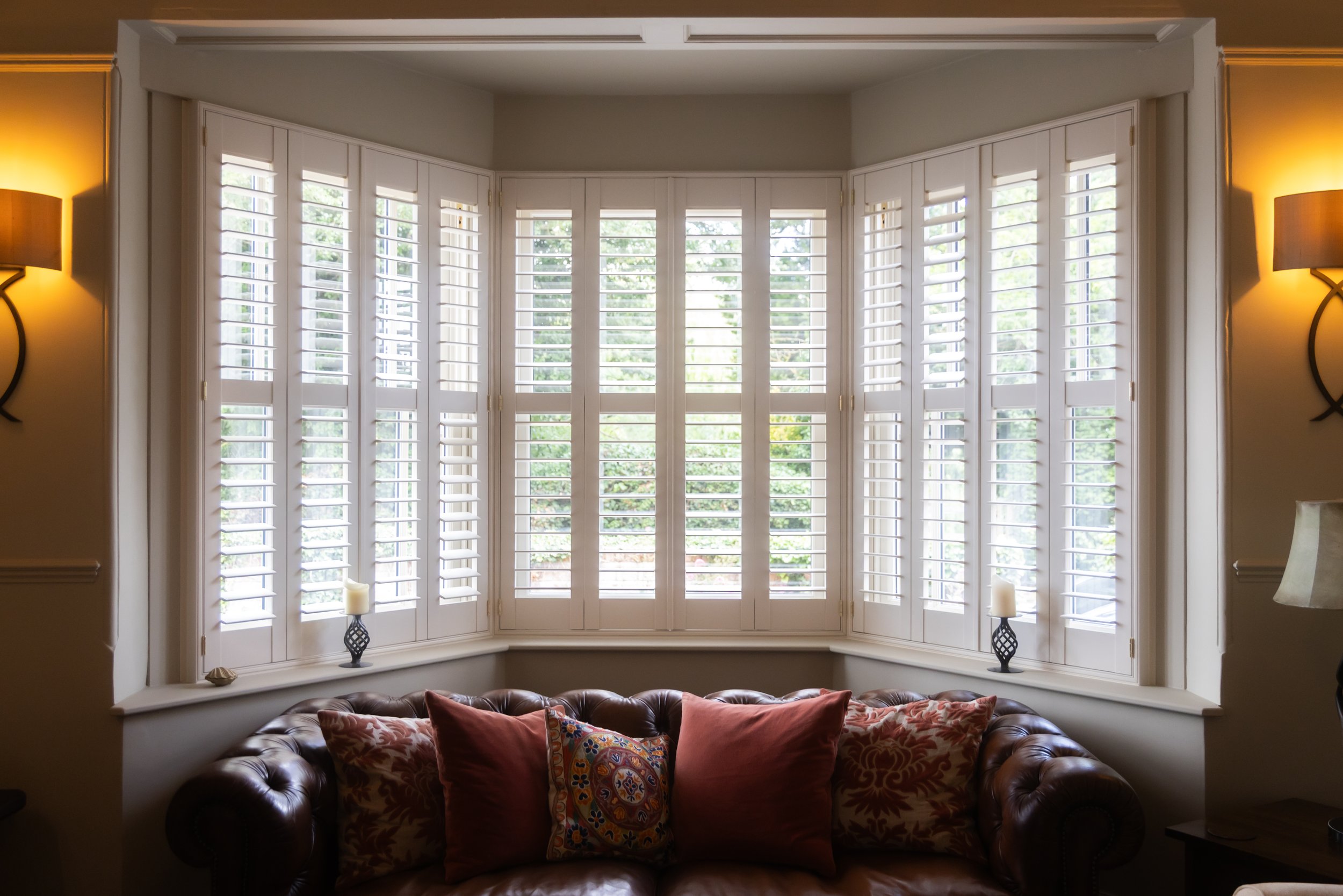 Living room with large bay window and white shutters, two wall sconces, and a dark leather sofa with colorful cushions.