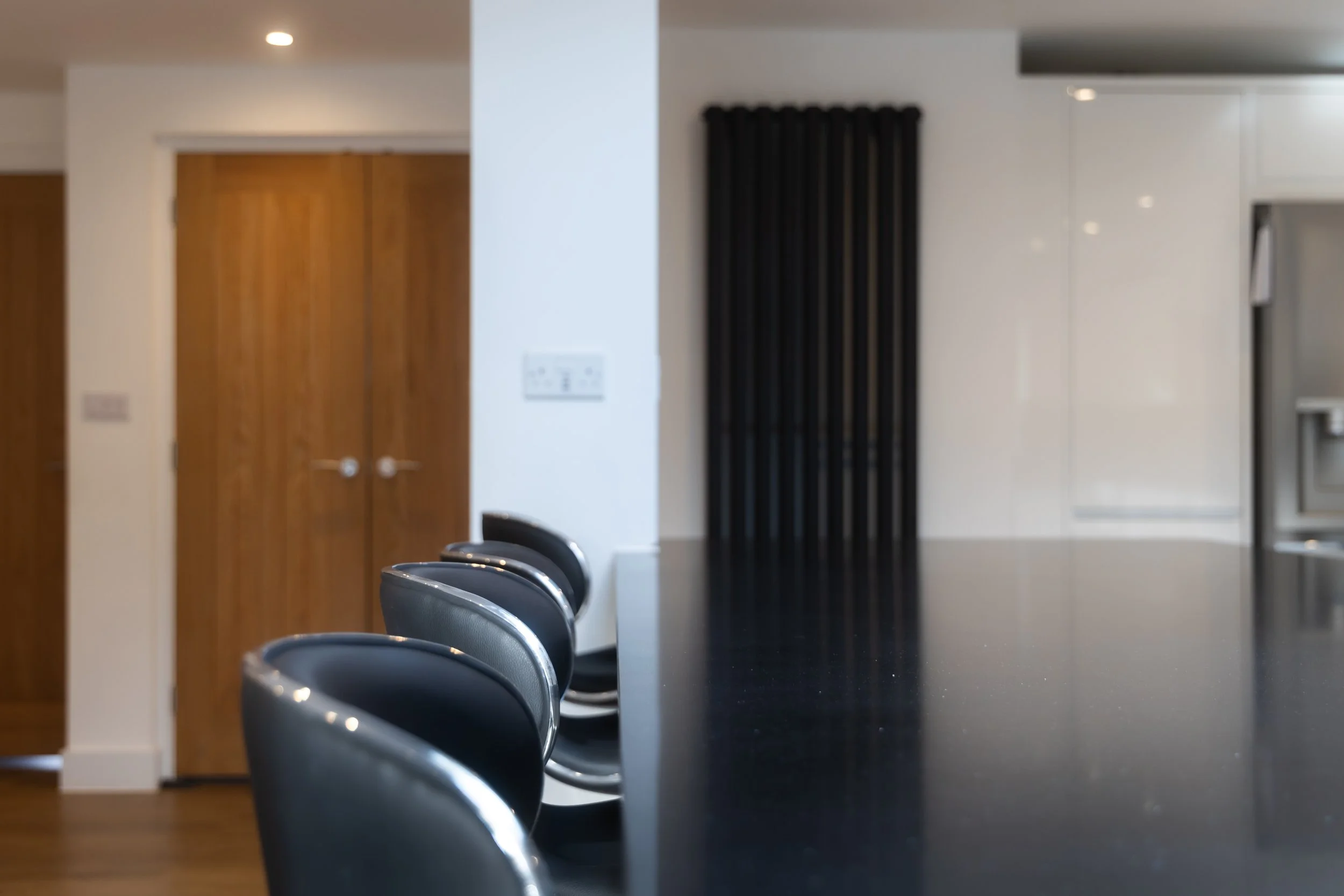 Interior of a modern room with black chairs lined up along a table, a wooden door on the left, a white wall with a control panel, and a window with black curtains. The space appears clean and minimalistic.