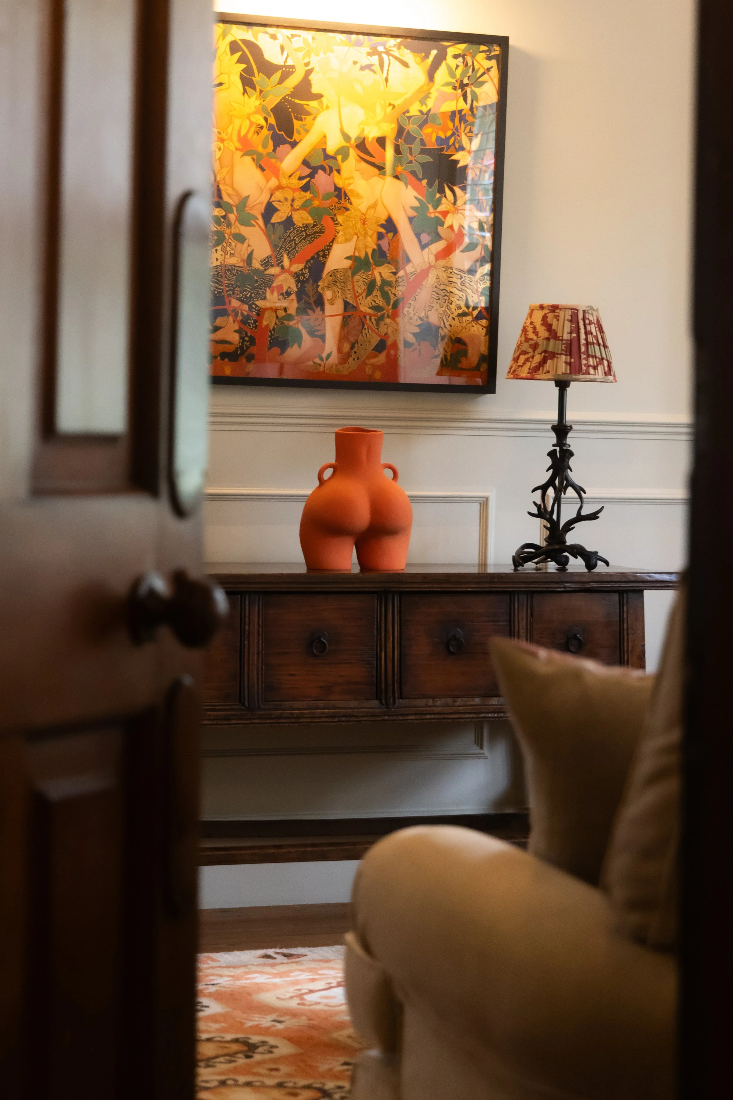 A view through an open door shows a wooden sideboard with a red vase, a table lamp with a branch-like base and a patterned shade, a framed artwork of a nude woman surrounded by leaves and animals, and part of a beige armchair with a pillow, in a well