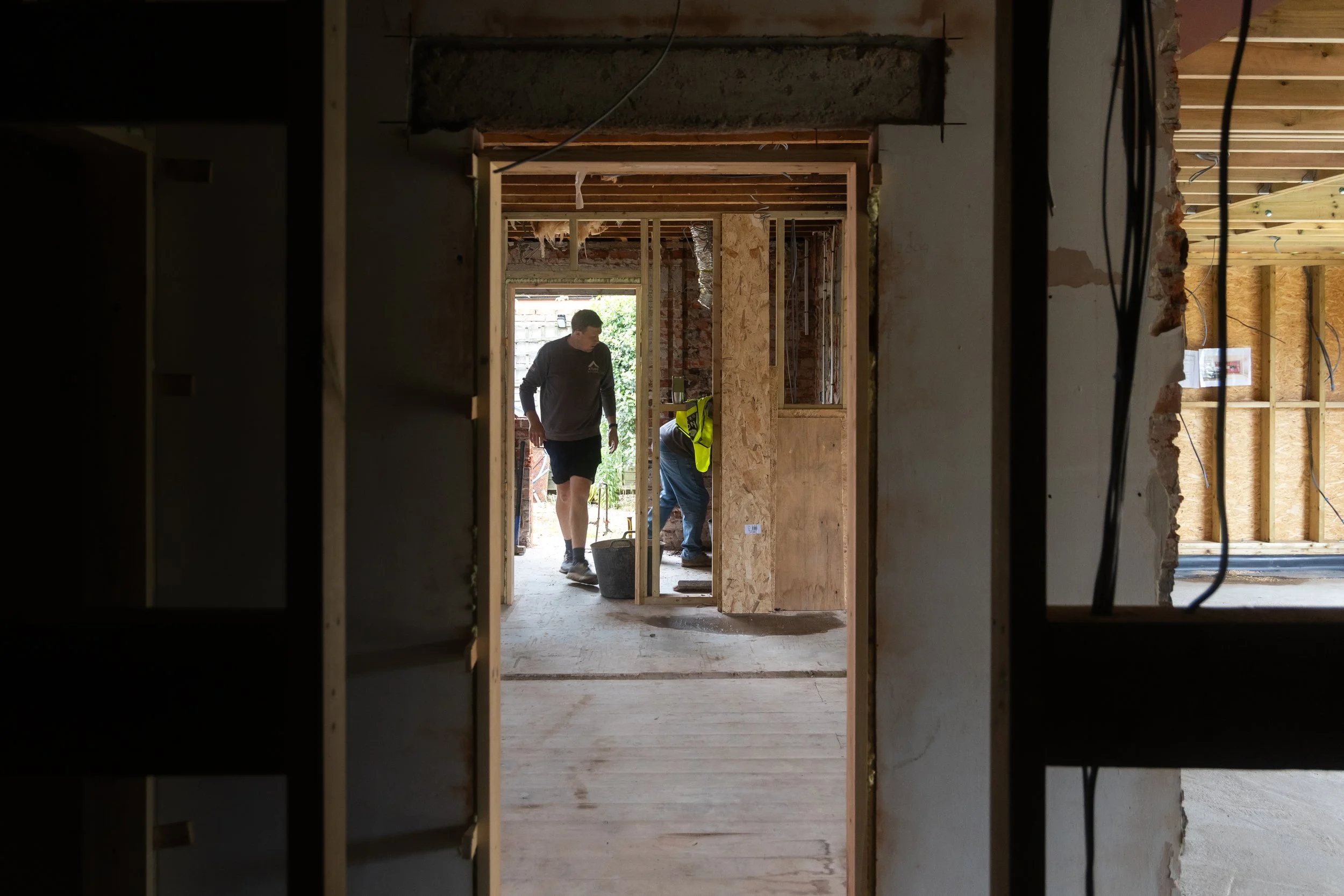 Construction workers inside a house under renovation, framing walls with wooden studs and exposed ceiling beams.