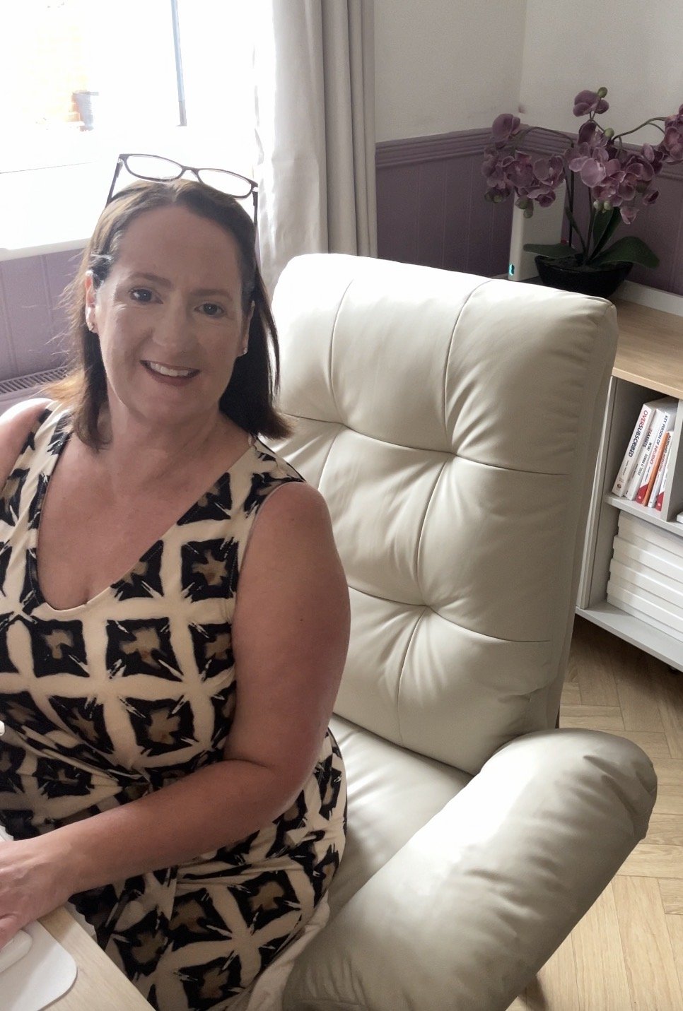 A woman with glasses on her head, wearing a patterned sleeveless dress, sitting on a white leather armchair in a bright room with a bookcase and a purple orchid plant in the background.