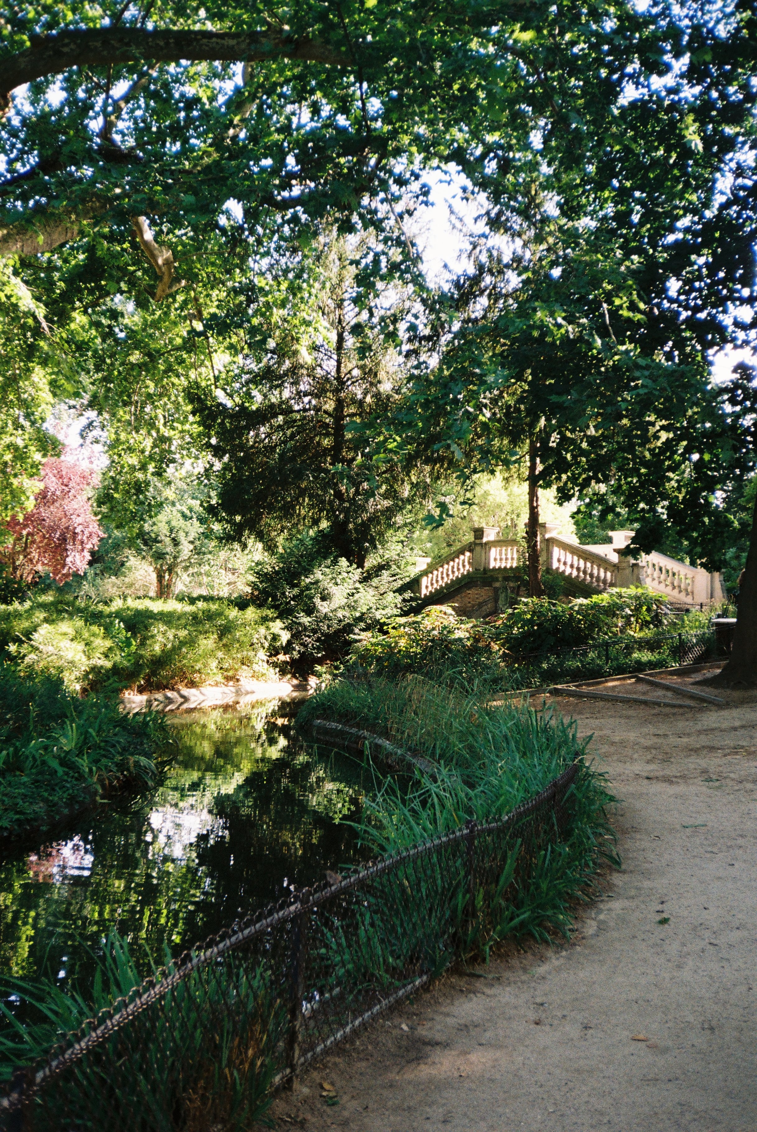 Ein grüner Park mit Bäumen, einer kleinen Wasserstraße, einem Weg und einer Steinterrasse mit Geländer.