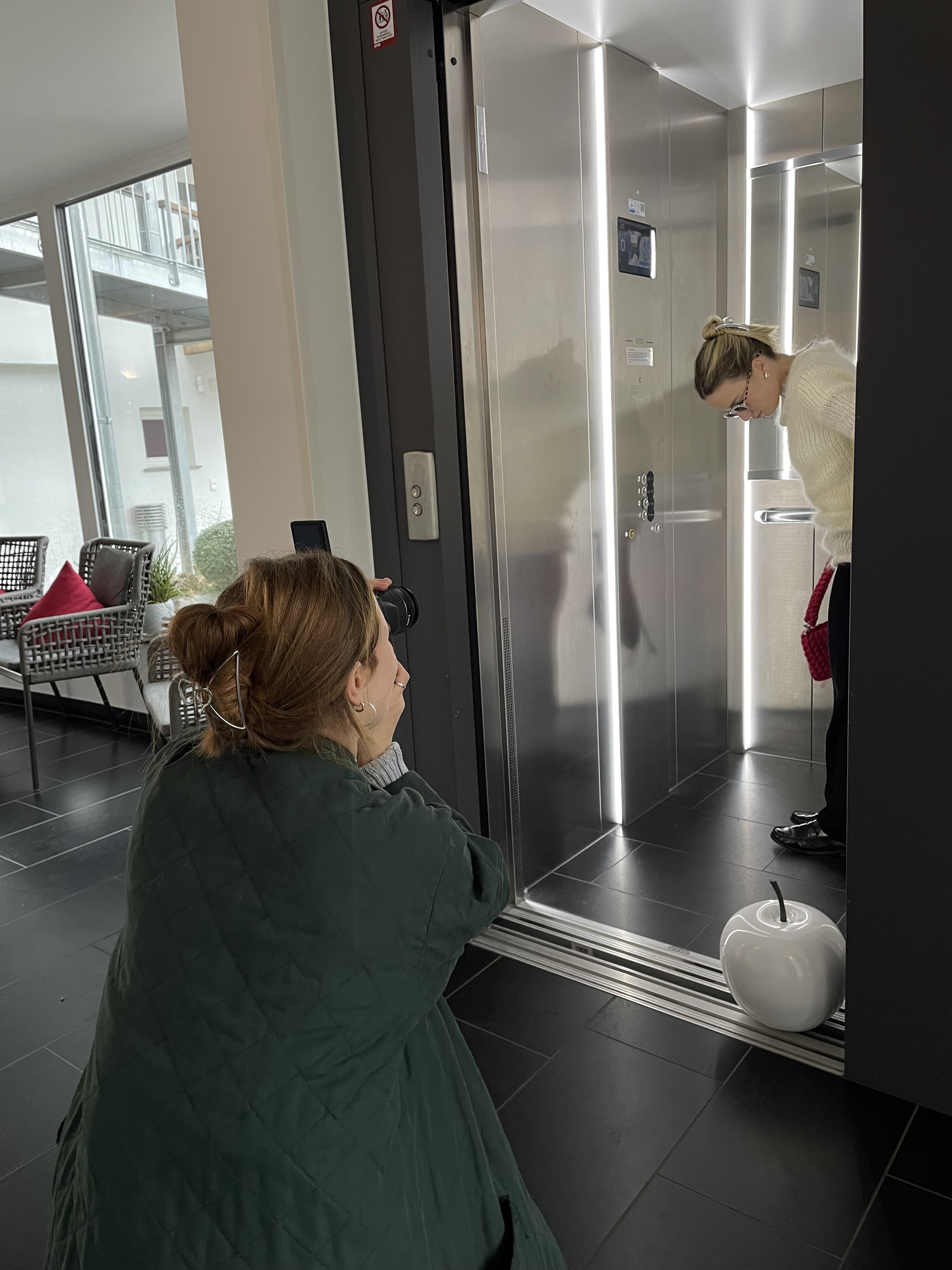 Zwei Frauen befinden sich in einem Aufzug, eine steht an der Tür, die andere sitzt in einem Wartebereich mit Stühlen und Kissen im Hintergrund.