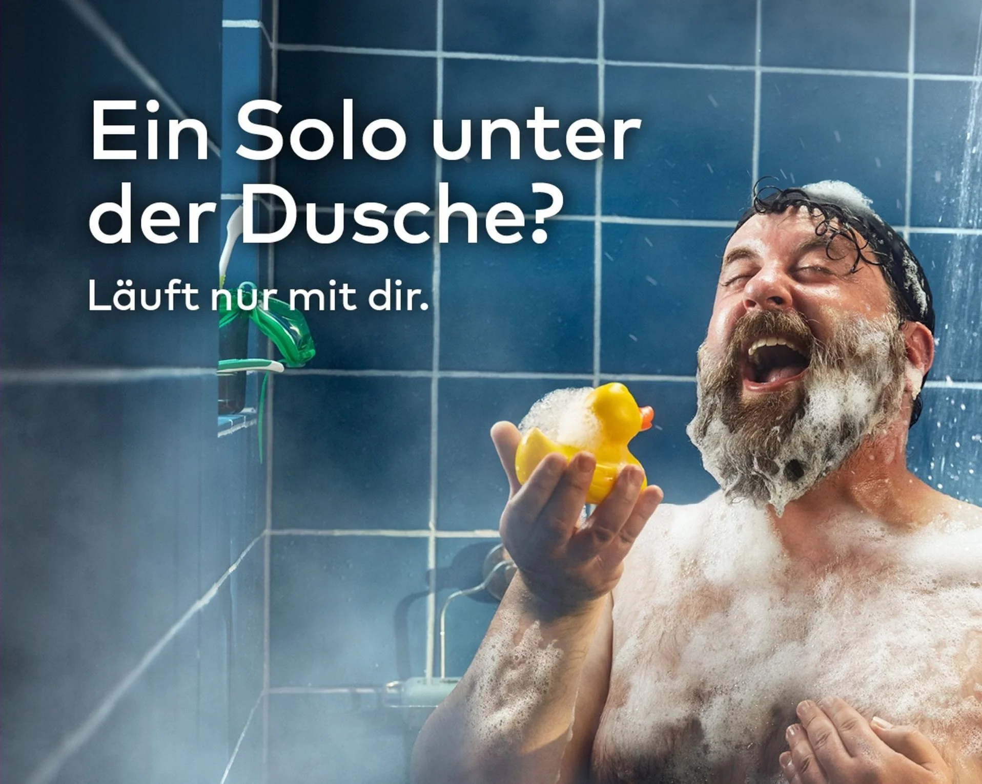 Ein Mann mit dunklem, nassem Haar und Bart, der in der Dusche steht, warmes Wasser läuft auf ihn. Er ist glücklich, lacht und hält eine gelbe Gummiente in der Hand. Die Wände sind blau mit weißen Fliesen, und es gibt eine kleine Ablage mit einer Zahnbürste an der Wand.