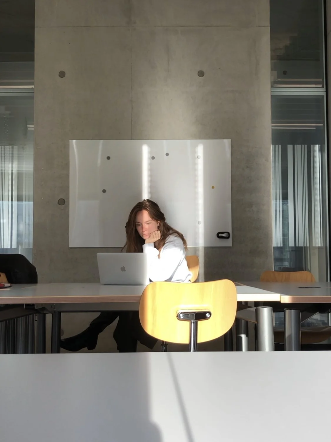 Junge Frau arbeitet an einem Laptop in einem modernen Büro, sitzend an einem Tisch mit mehreren Stühlen. Helles Licht fällt durch Fenster, und eine weiße Tafel hängt an der Wand im Hintergrund.