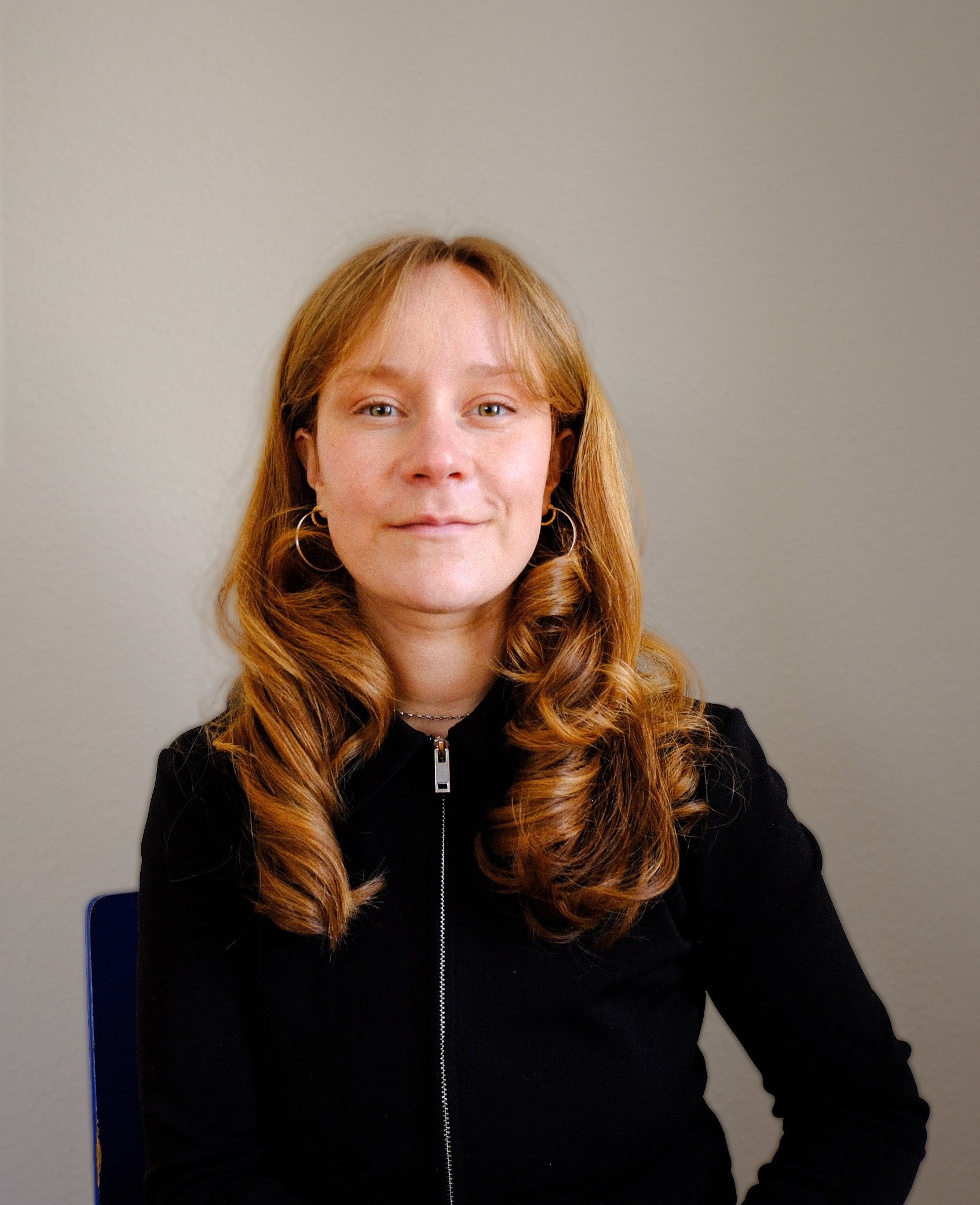 Junge Frau mit roten, lockigen Haaren und goldenen Ohrringen, sitzt auf einem blauen Stuhl.