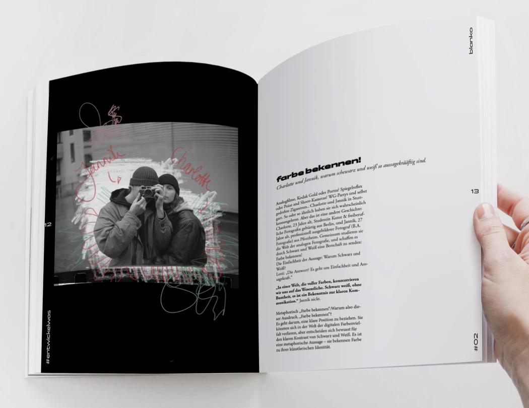 Eine Hand hält ein Magazin mit einer schwarzen und weißen Fotoseite, auf der ein Foto von zwei Menschen, die sich fotografieren, mit bunten Kritzeleien und Namen darüber zu sehen sind. Der Text auf der rechten Seite handelt von Farben.