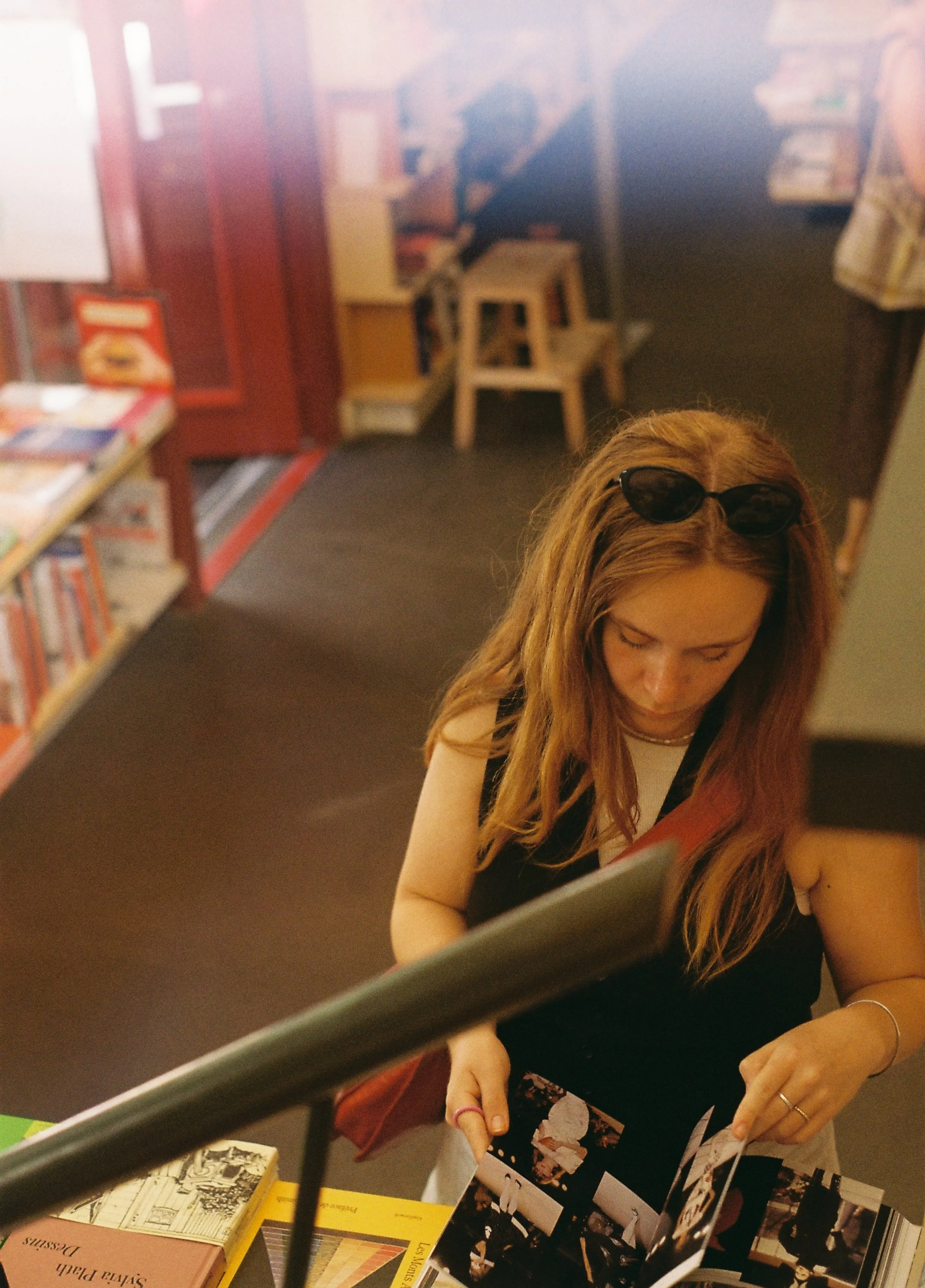 Eine junge Frau mit langen roten Haaren, Sonnenbrille auf dem Kopf, blättert durch eine Fotobroschüre in einem Buchladen oder einer Buchhandlung.