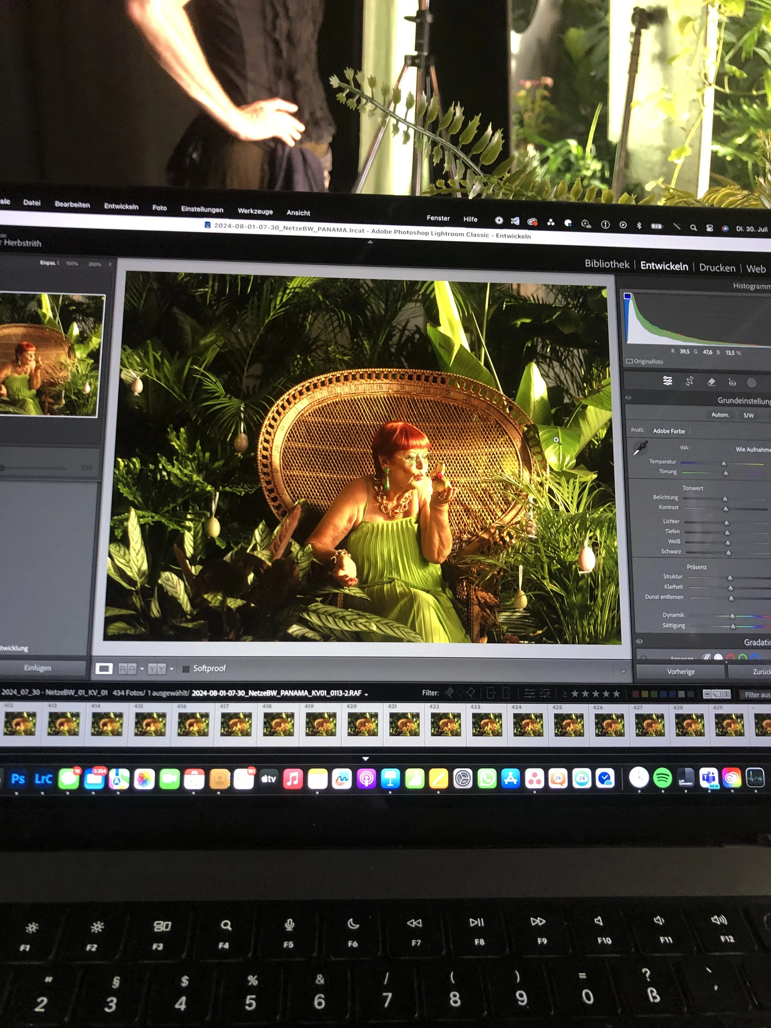 Ein Laptop-Bildschirm zeigt eine Fotobearbeitung mit einer Frau, die in einem grünen Kleid auf einem Stuhl sitzt, umgeben von Pflanzen. Es ist eine Aufnahme im Nachtmodus, mit sichtbaren Bearbeitungswerkzeugen und Histogrammen auf der rechten Seite.