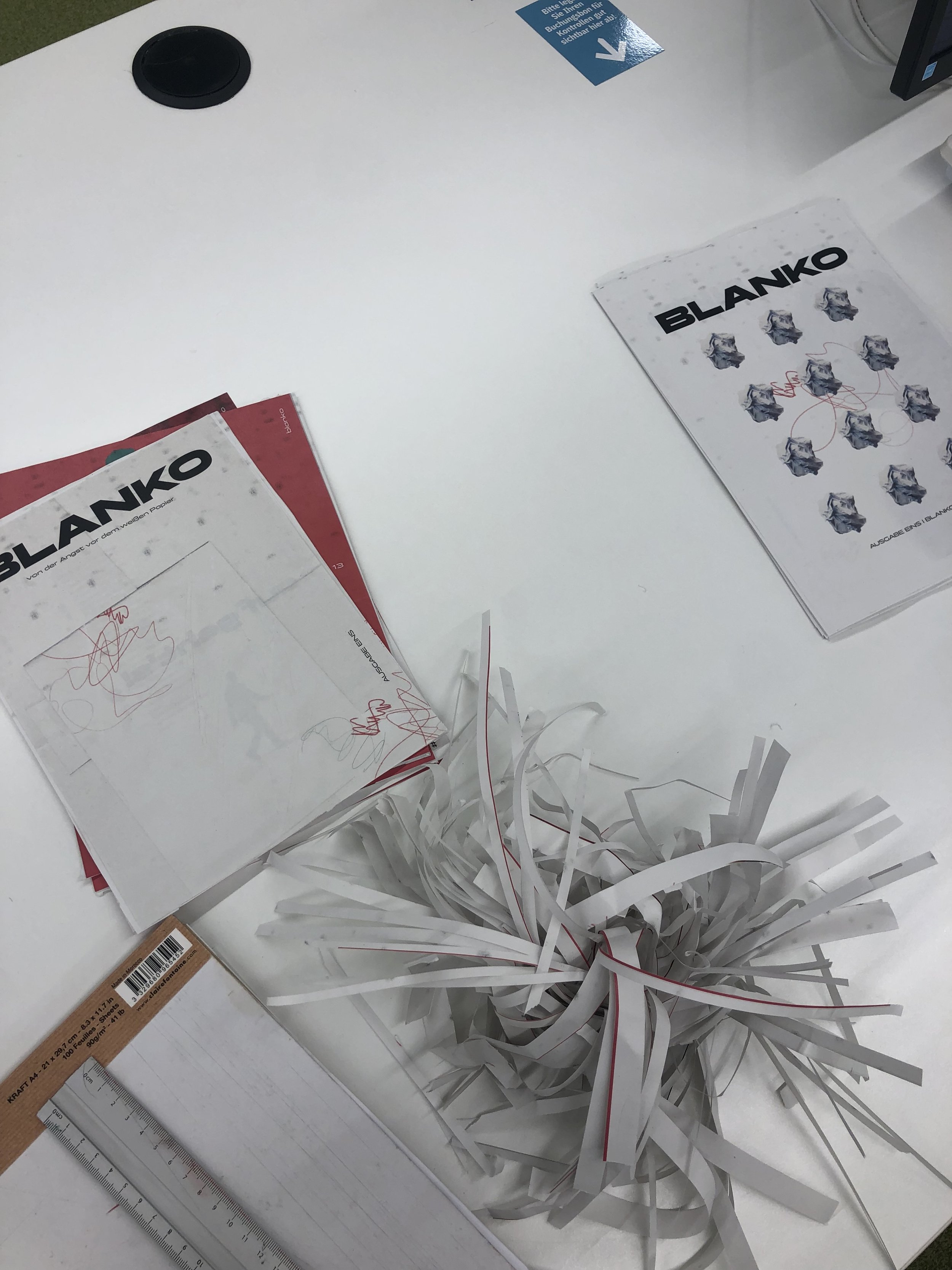 Auf einem weißen Tisch liegen mehrere Papiere, ein Lineal, Papierstreifen und ein Teller. Das Papier trägt den großen Schriftzug 'BLANKO' und zeigt bunte Bilder von zerknülltem Papier. Es gibt auch eine Ecke mit einem blauen Sticker, der Anweisungen auf Deutsch enthält.