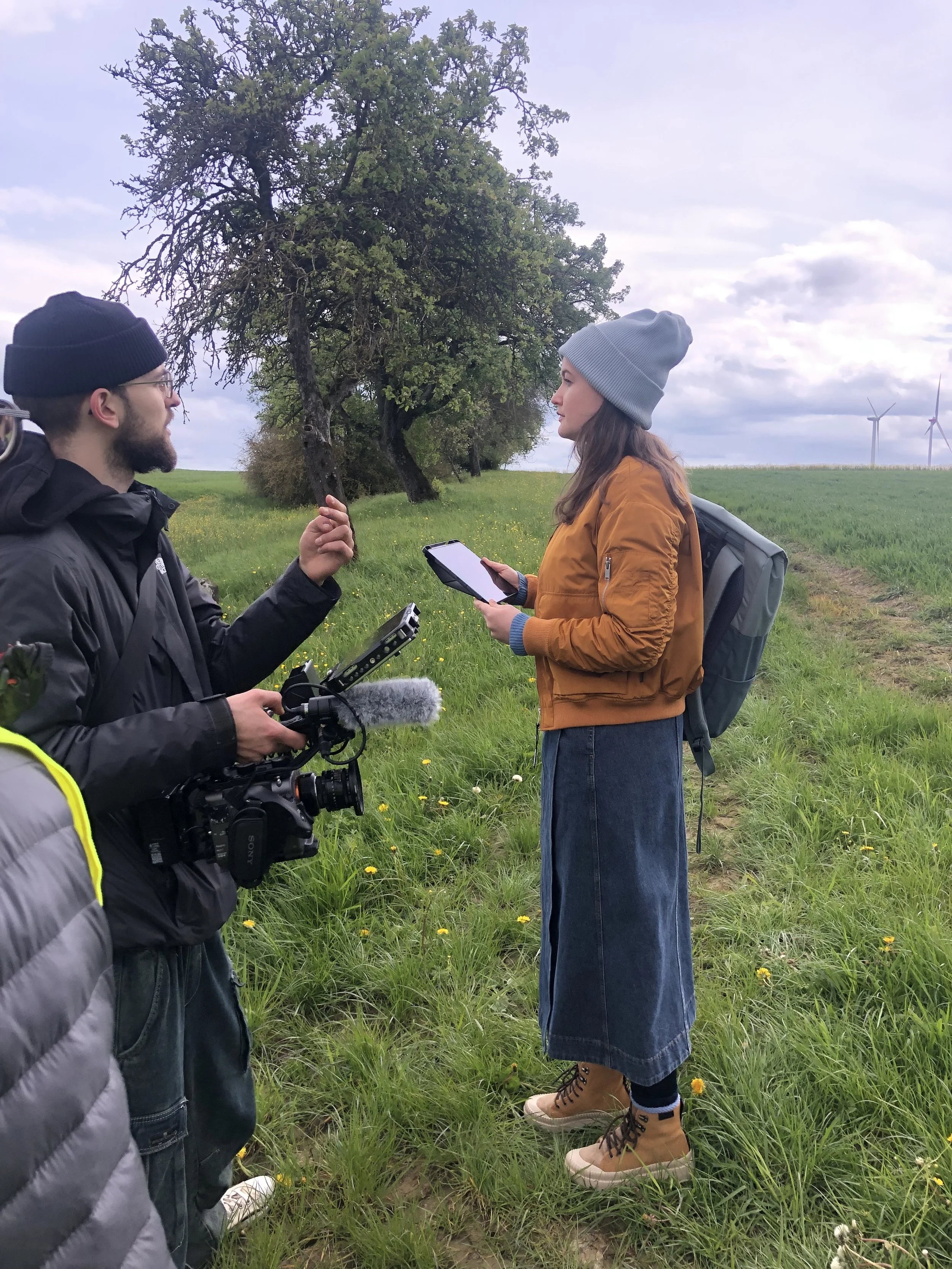 Zwei Personen in einem Feld, die sich unterhalten. Im Hintergrund stehen Bäume und Windkraftanlagen. Es ist bewölkt.