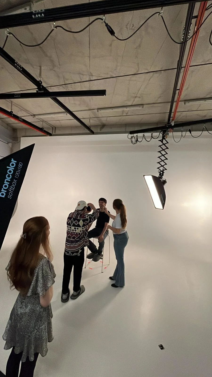 Menschen im Fotostudio, eine Person sitzt auf einem Hocker und lässt sich fotografieren, während drei andere Frauen um sie herumstehen, eine hält das Handy für das Foto, die andere spricht mit ihm, im Hintergrund sind Studioausrüstung und ein Beleuchtungsgerät sichtbar.