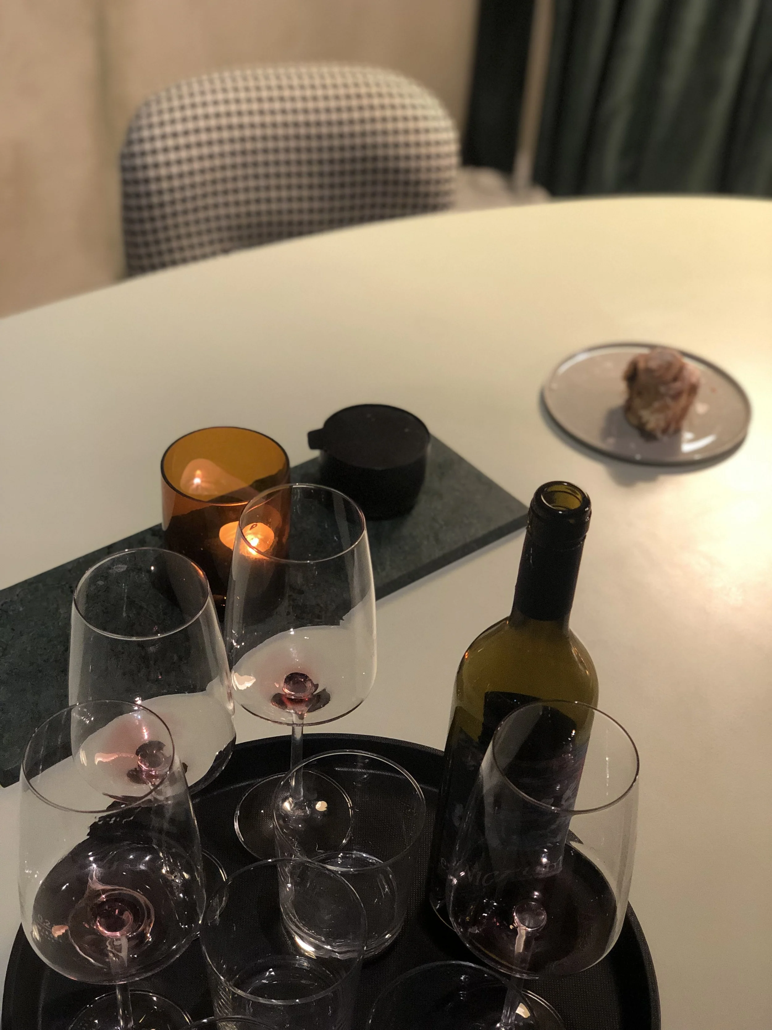 Blick auf mehrere leere Weingläser auf einem schwarzen Teller mit einer Flasche Wein im Vordergrund, im Hintergrund eine Kerze, ein Teller mit Fleisch und ein Stuhl.