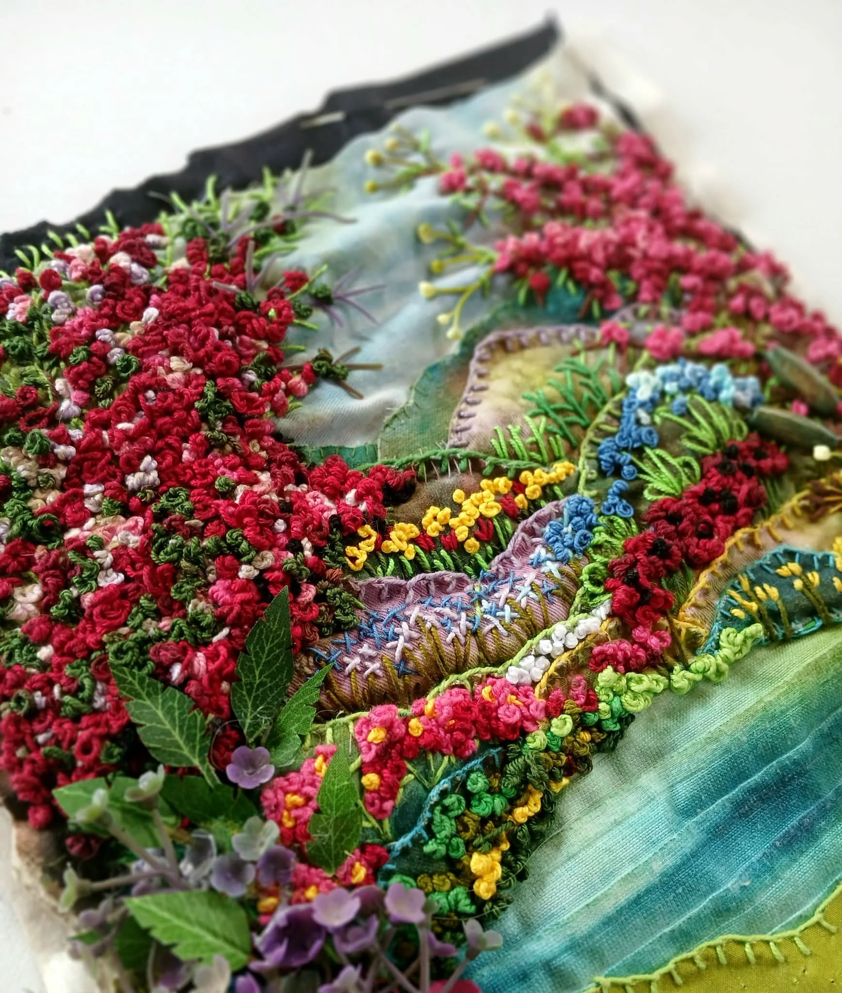 Close-up of embroidered fabric with colorful floral patterns, placed on layered textured cloths in shades of blue and green, with some green leaves and purple flowers in the foreground.