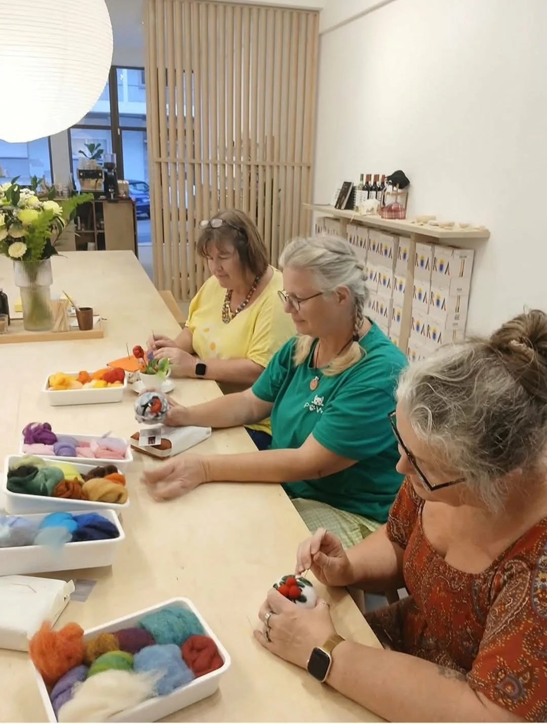 Four women sitting at a light wood table, crafting with colorful felt and stuffing, surrounded by supplies including bowls of felted wool, in a well-lit room with a vase of flowers and a paper lantern.