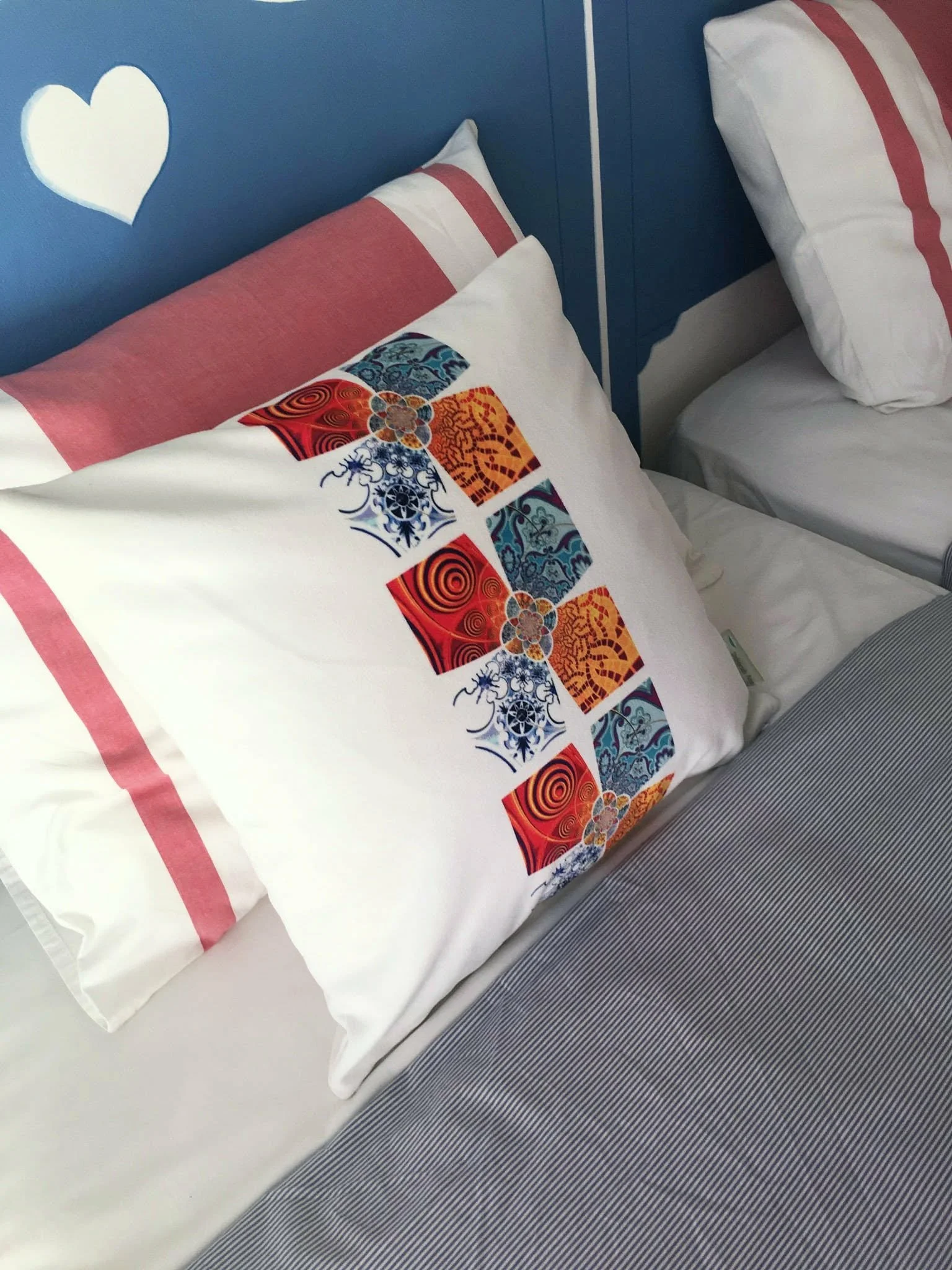 A close-up of two beds with white pillows, one with a decorative pillow featuring colorful, intricate pattern, inspired by the tiles of Tavira. Available in 3 different design and 6 different colourways.