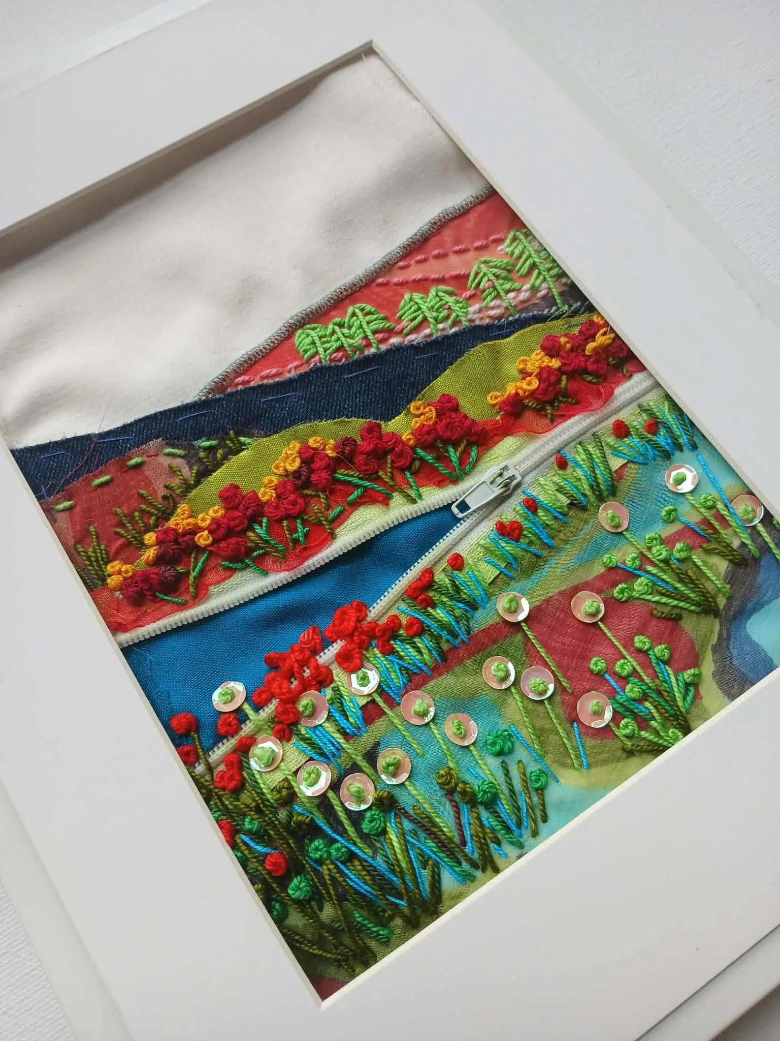 Close-up of colorful embroidery work on fabric with sequins and various stitch patterns in a white frame.