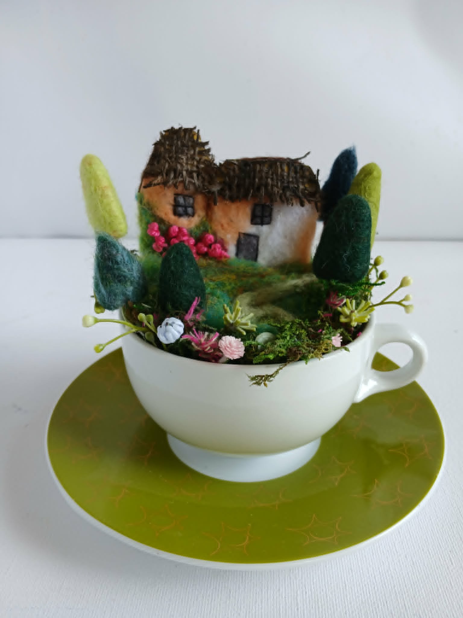 A decorative miniature house with a thatched roof, surrounded by felt trees, flowers, and foliage inside a white teacup on a gold plate.