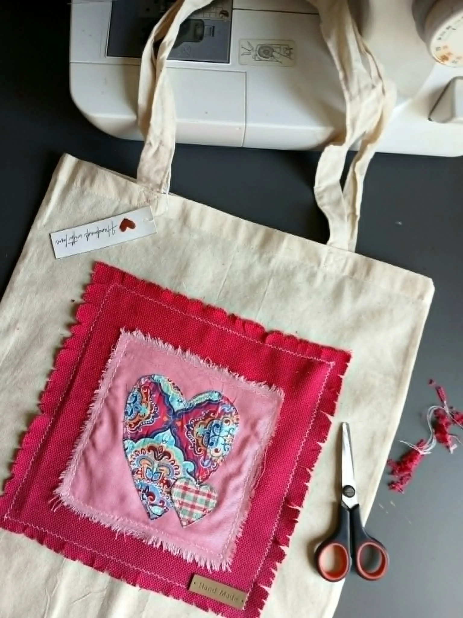 An eco linen tote with a layered handmade heart design, on the front of the bag, accompanied by a pair of black and orange scissors, and a small rectangular tag that says "handmade with love".