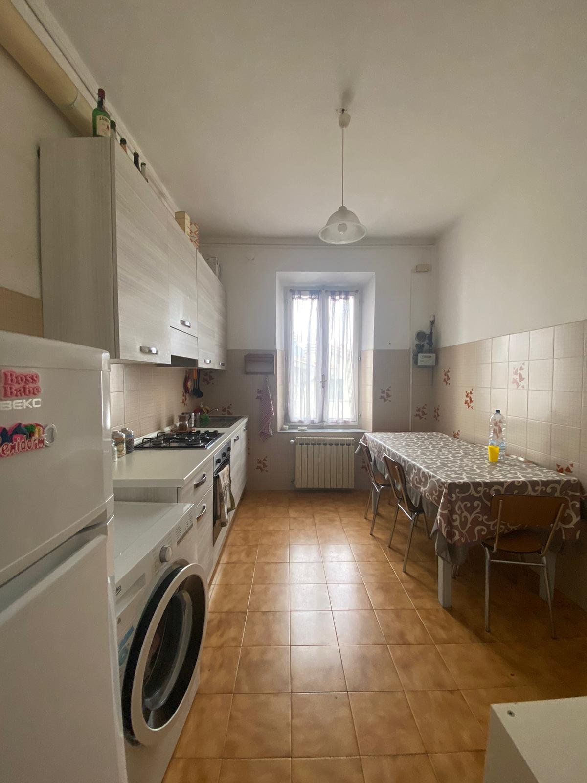 Cucina con mobili bianchi, tavolo con tovaglia decorata, frigorifero, forno, piano cottura, un divano sotto una finestra, pareti rivestite di piastrelle beige con motivi floreali.