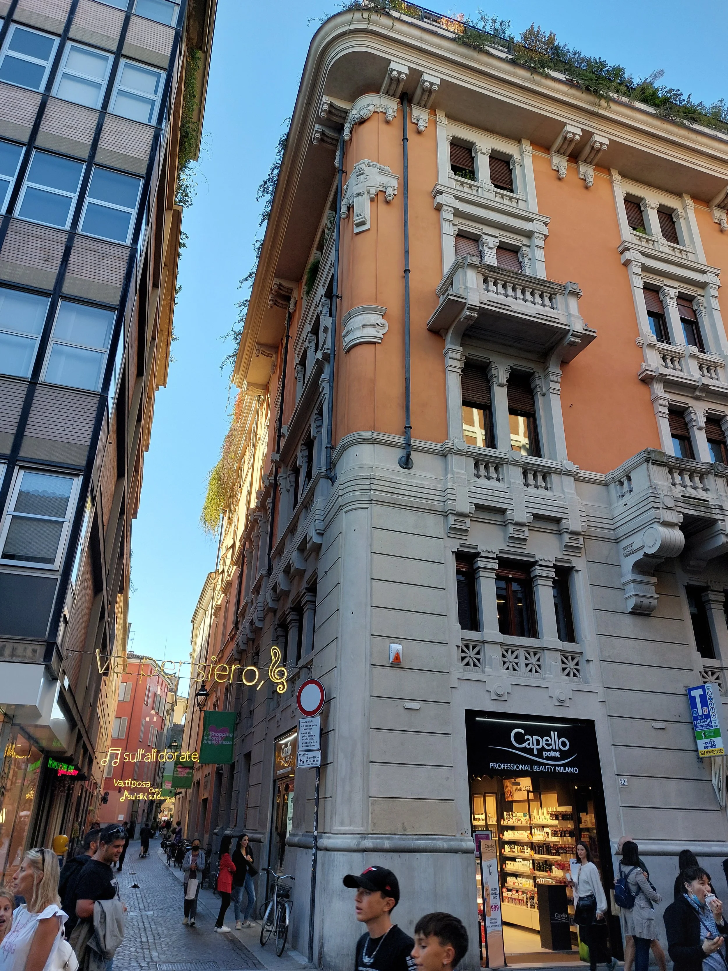 Via affollata con negozi, persone che camminano, edifici vecchi e moderni, cielo azzurro, negozio di prodotti di bellezza chiamato Capello Point
