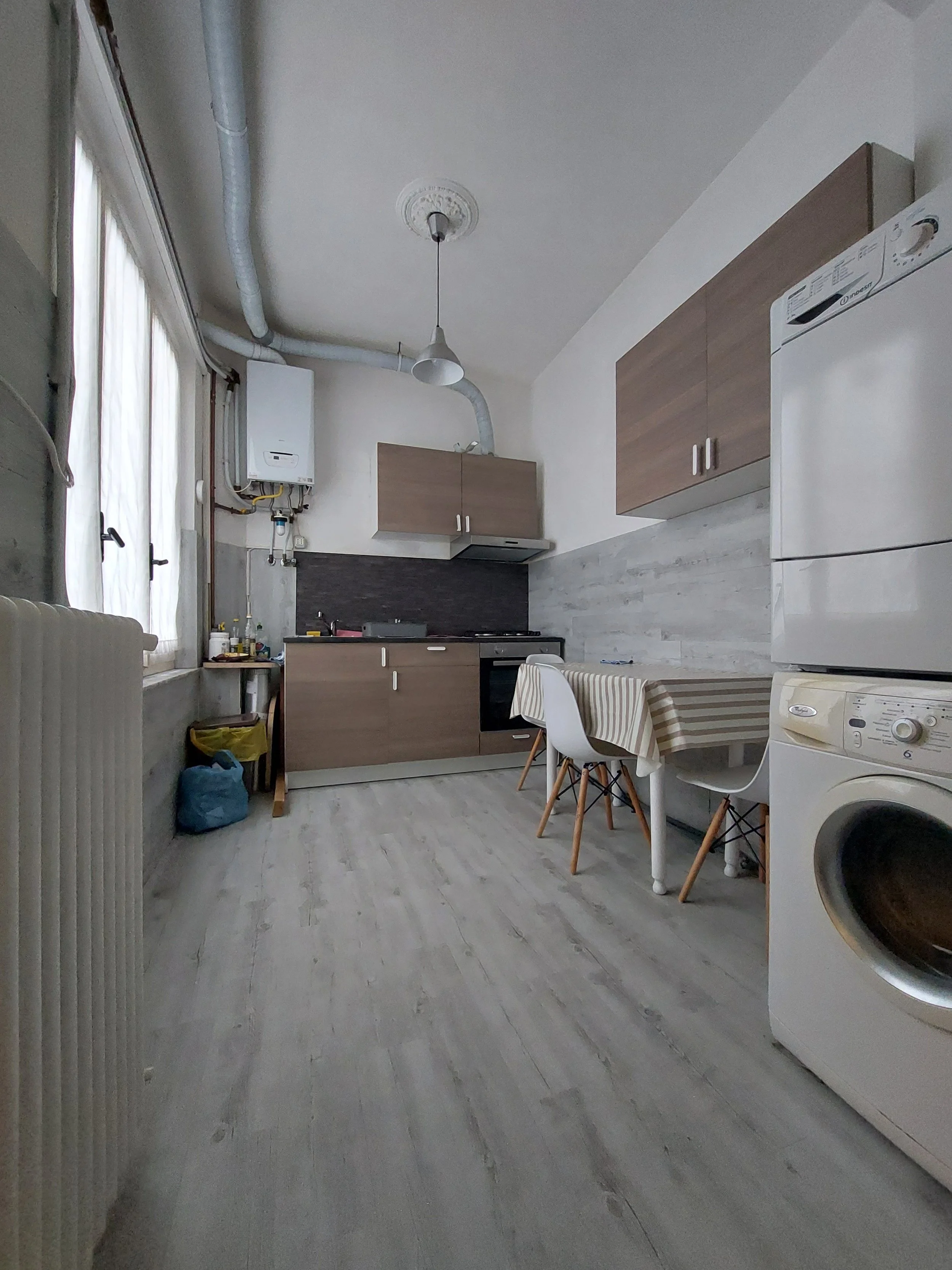 Una cucina moderna con mobili in legno, un tavolo tondo coperto con una tovaglia a righe, sedie di design e un elettrodomestico bianco, presenza di una finestra con tende leggere e una parete con dettagli di pietra.