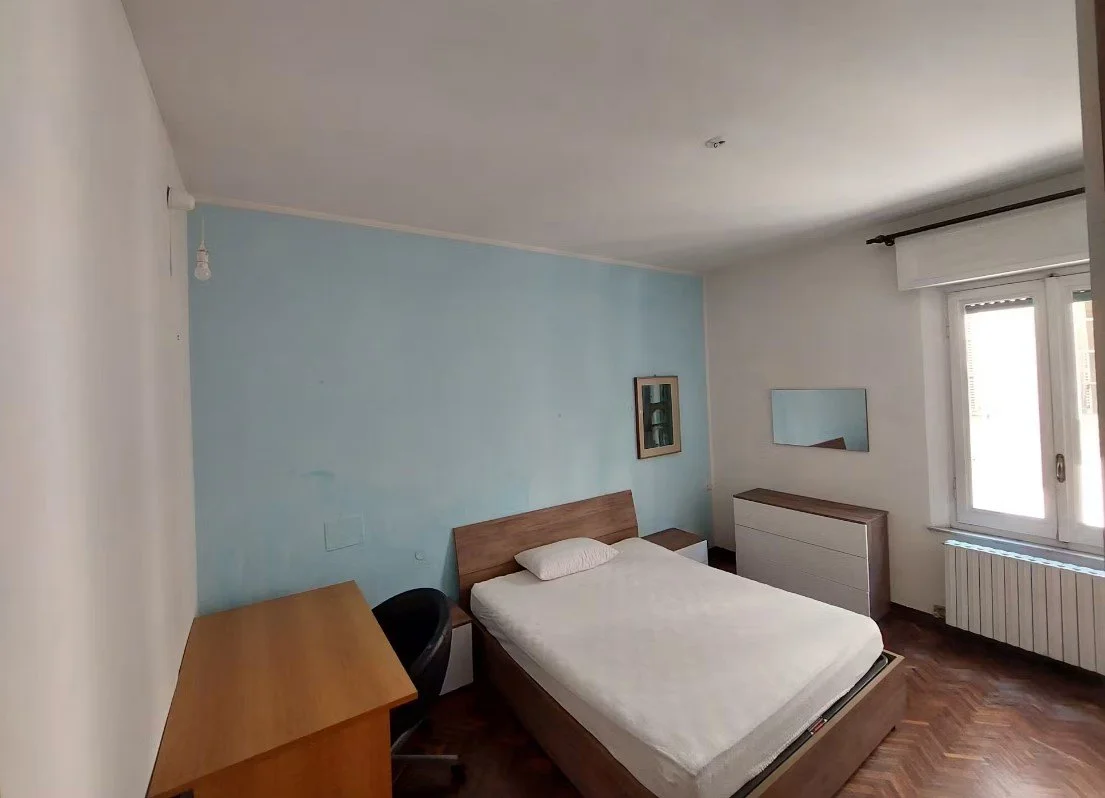 Camera da letto semplice con letto matrimoniale, armadio, scrivania, sedia, e un mobile con specchio vicino alla finestra.