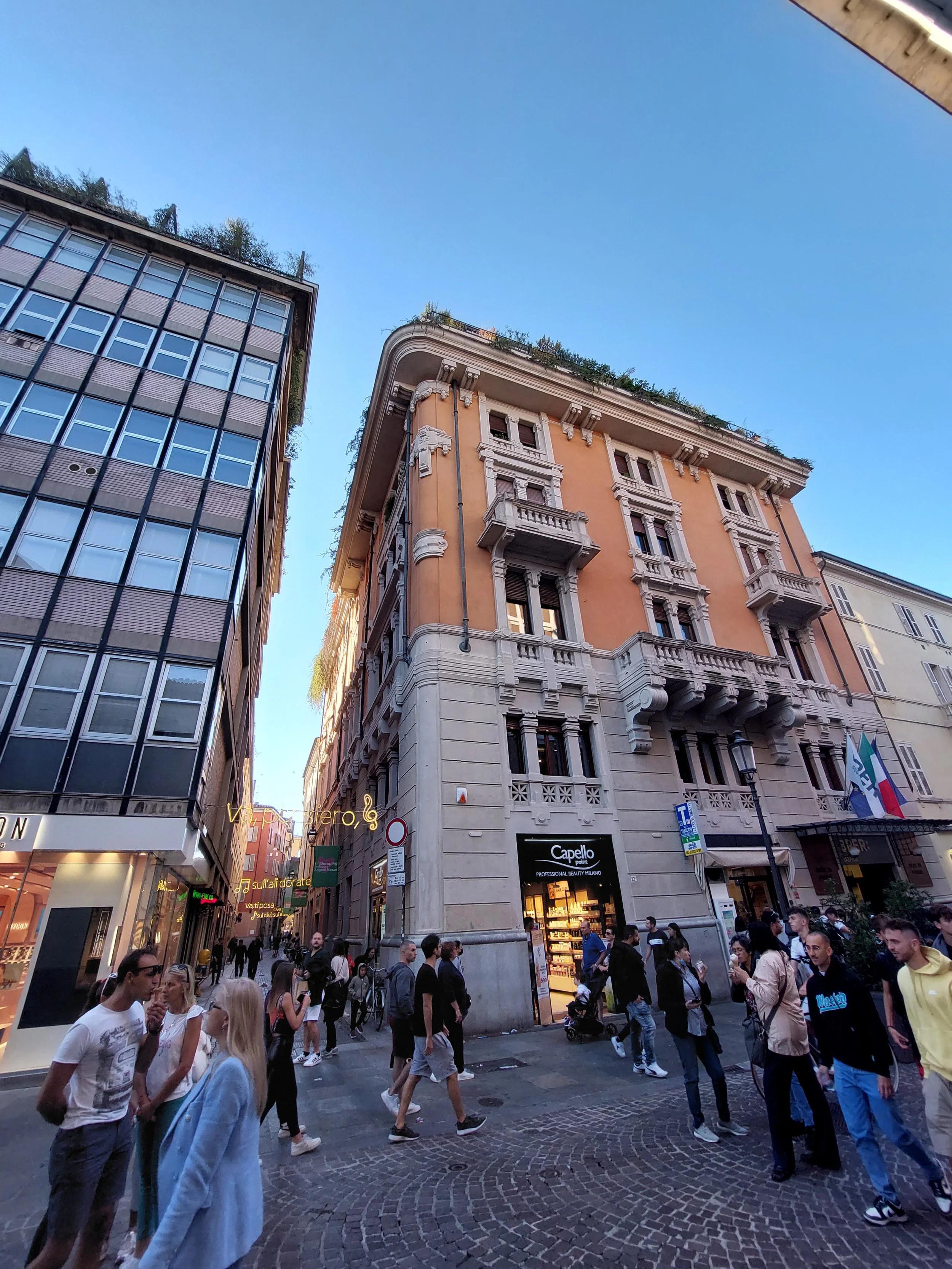 Vista di una strada affollata con persone che camminano davanti a negozi e edifici storici, sotto un cielo azzurro in una giornata di sera a Torino, Italia.