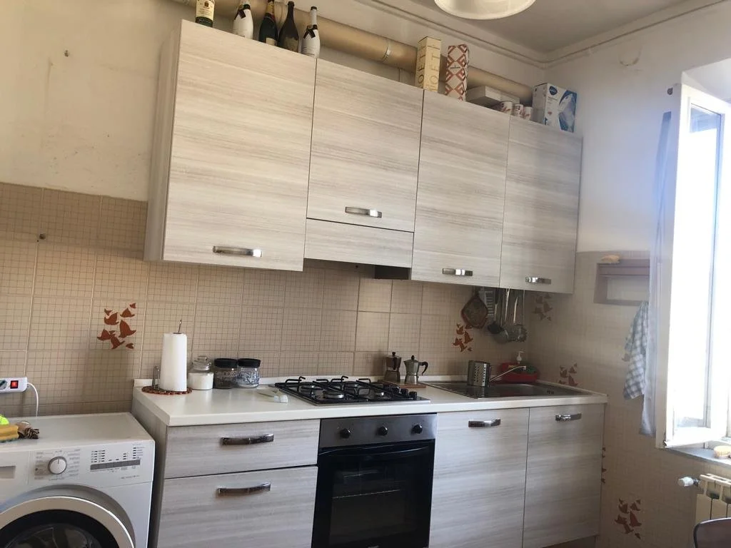 Cucina con armadietti superiori e inferiori in legno chiaro, piano cottura a quattro fuochi, forno, lavello, una finestra aperta a destra, impianto di riscaldamento, e vari oggetti sulla mensola e sul piano.