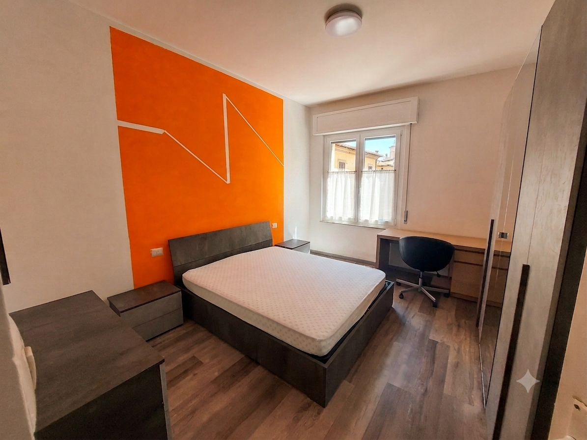 Camera da letto con parete orange, letto con materasso, due comodini, scrivania con sedia, armadio e finestra.