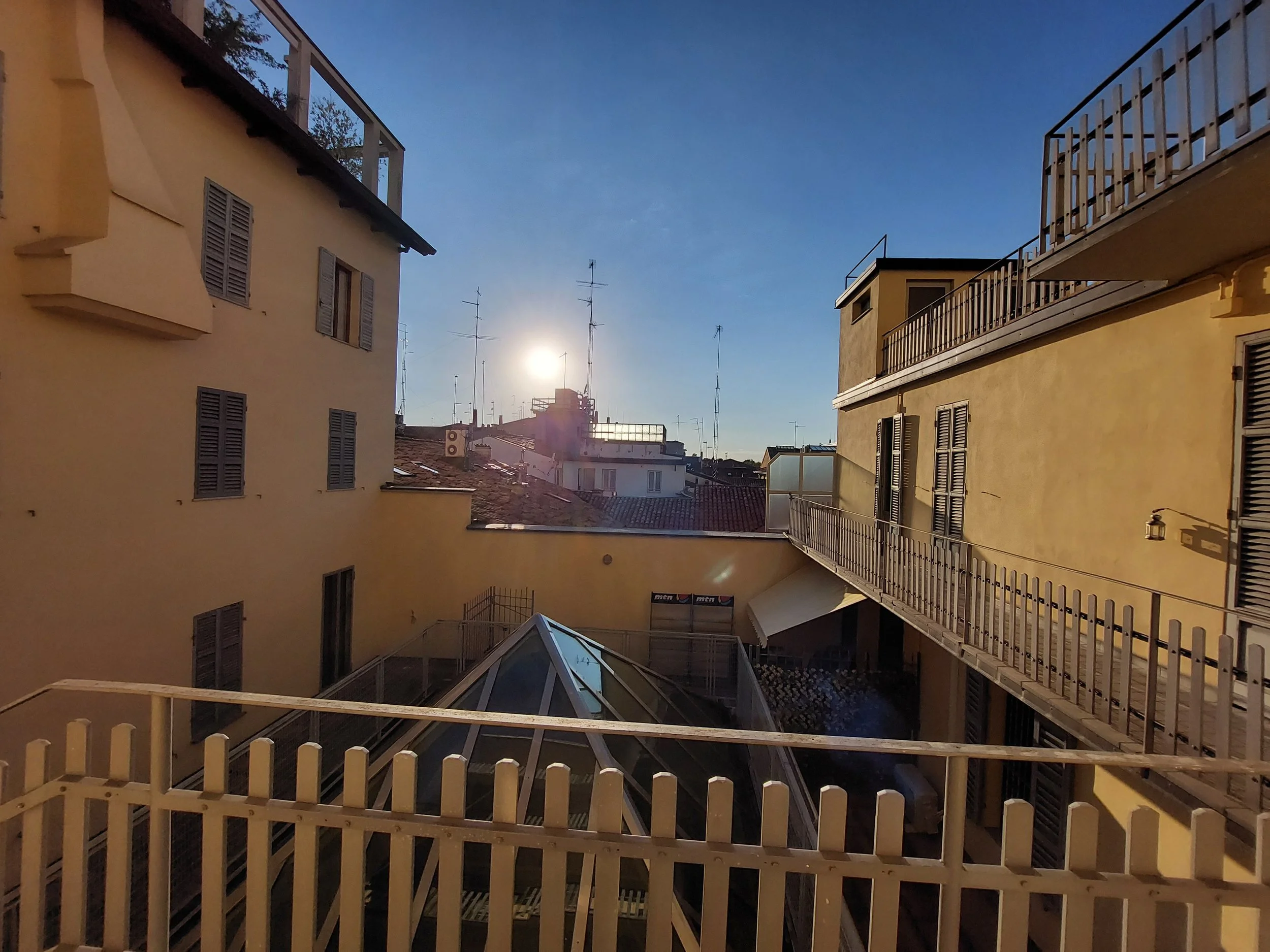 Veduta di un cortile di città con edifici gialli, balconi e antenne sul tetto, sotto un cielo sereno con il sole basso.