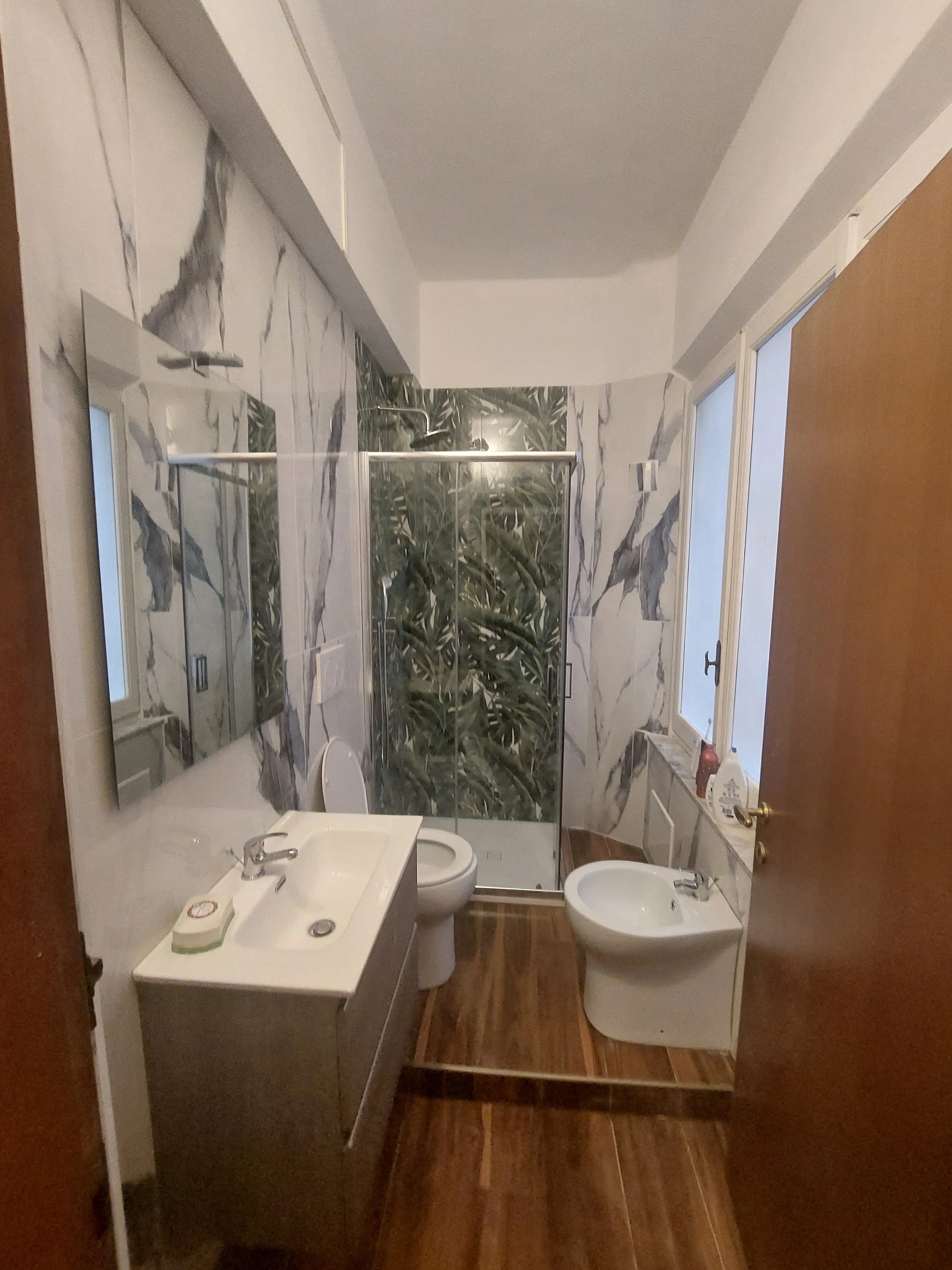 Bagno con pareti in marmo, doccia con vetri, lavabo, specchio, WC, bidet e porta in legno.