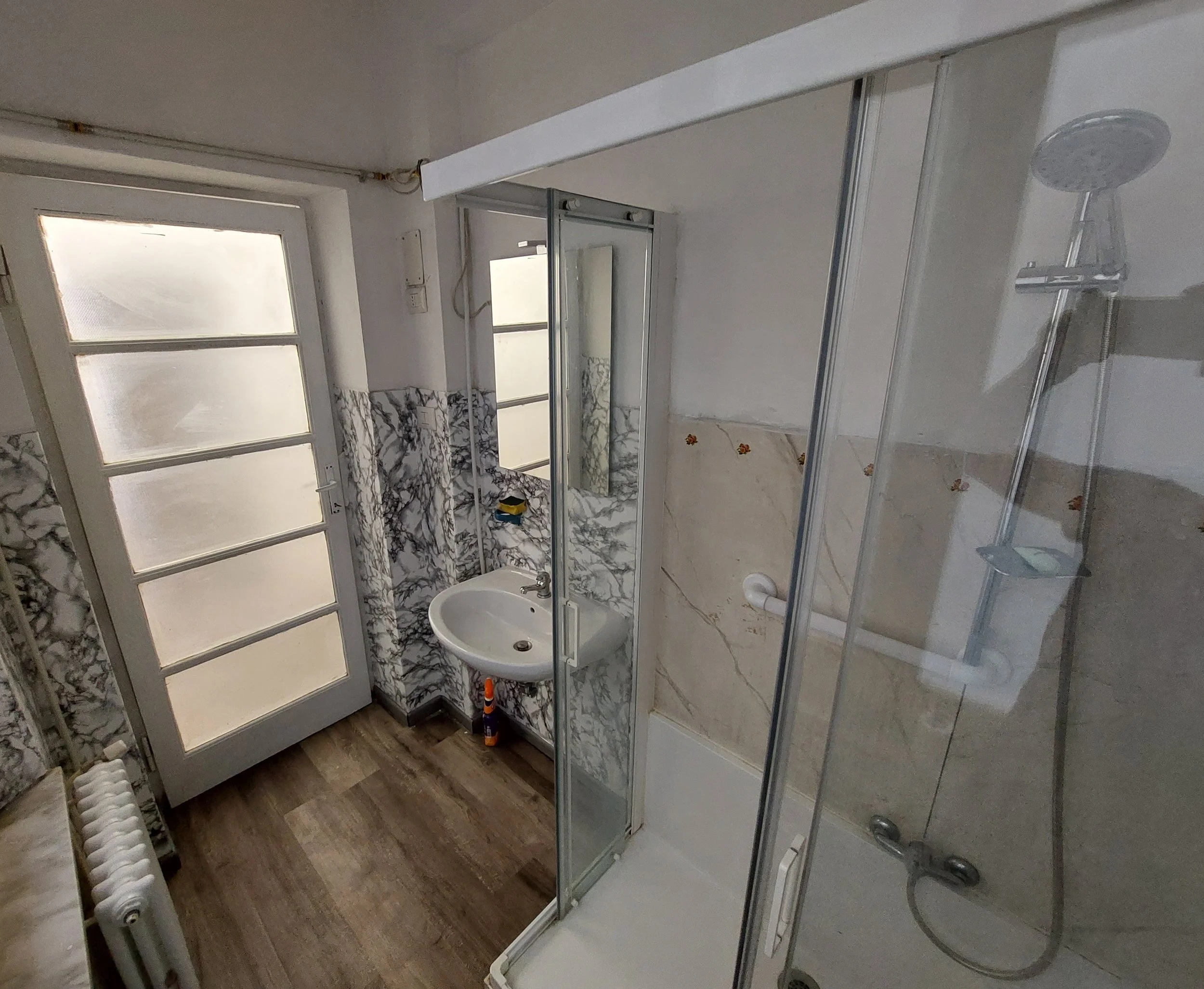 Interno di un bagno con doccia, lavabo e finestra, pareti rivestite con marmo e pavimento in legno.