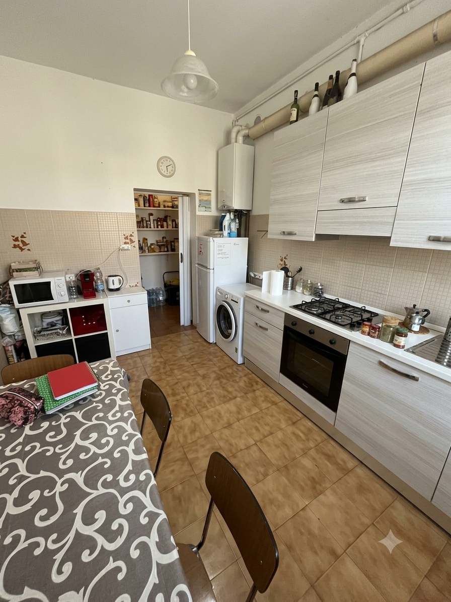 Cucina con mobili bianchi, forno, piano cottura a gas, lavatrice, frigorifero, disponibile anche una libreria con vari oggetti come bottiglie e scatole.