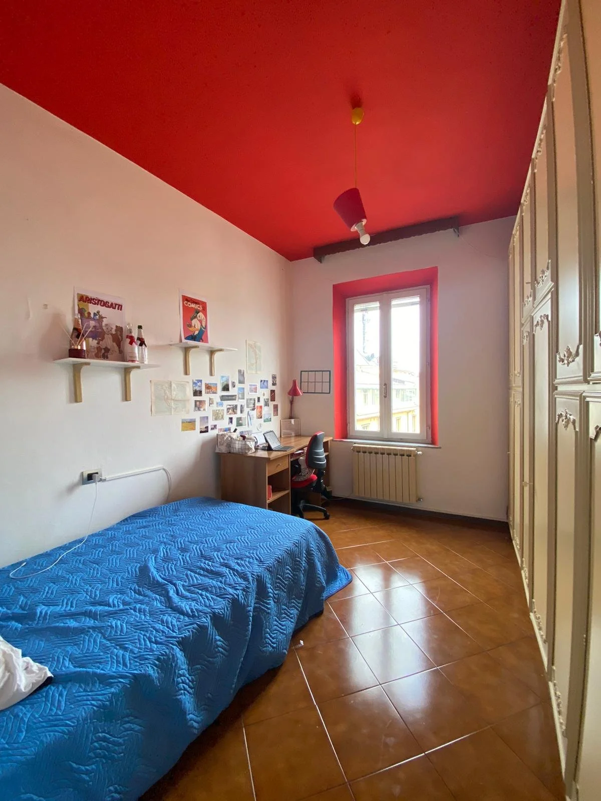 Una camera da letto con pareti bianche, soffitto rosso, un letto con copripiumino blu, una scrivania con sedia e una grande armadio bianca con dettagli decorativi, una finestra con cornice rossa, decorazioni sulla parete e una lampada da tavolo rossa.