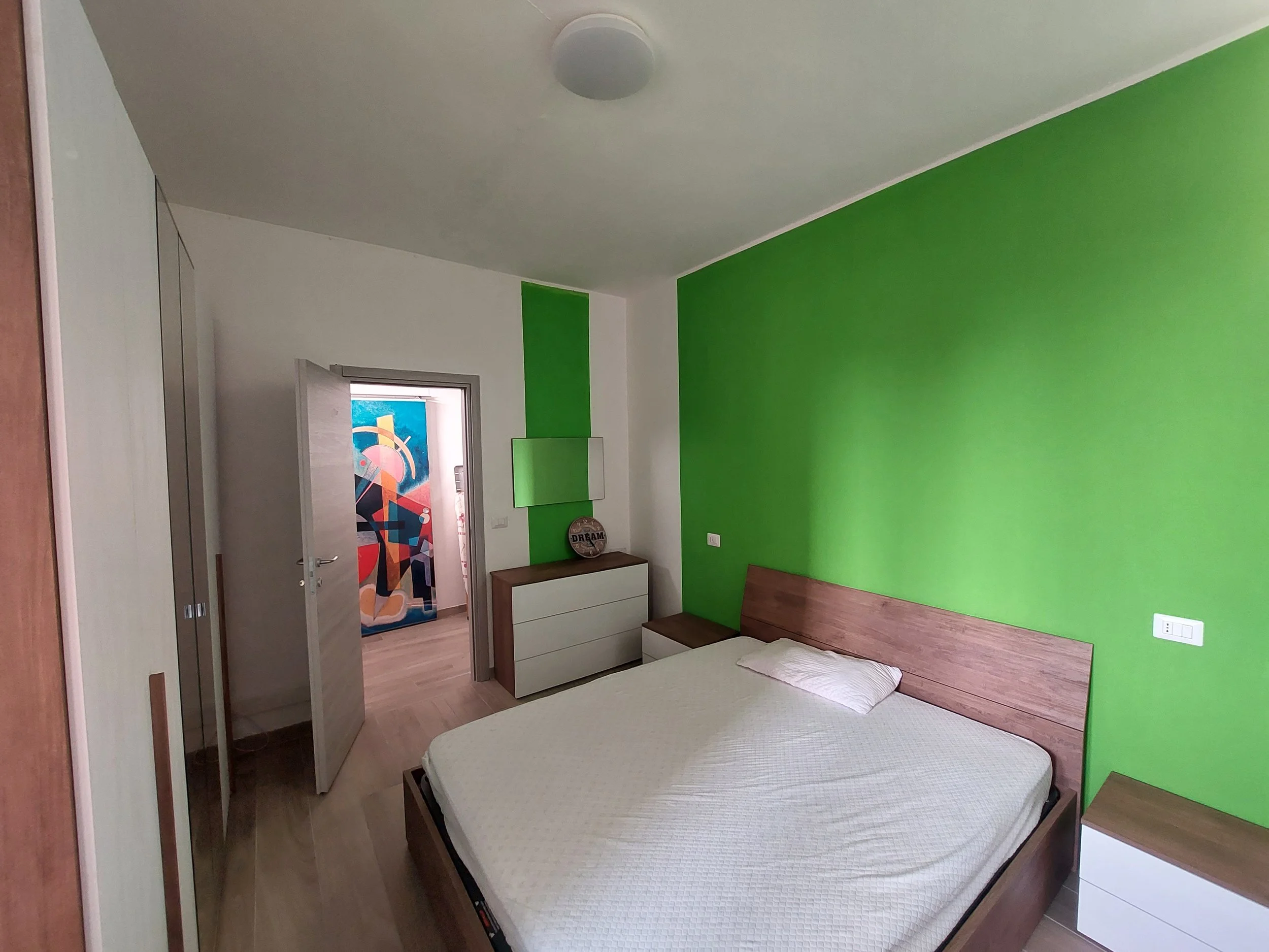 Camera che mostra una stanza con pareti di colore verde e bianco, un letto con coperta beige e un comodino in legno. Nell'angolo c'è un armadio chiuso e una porta con un quadro colorato appeso dietro. La stanza ha un pavimento di legno.