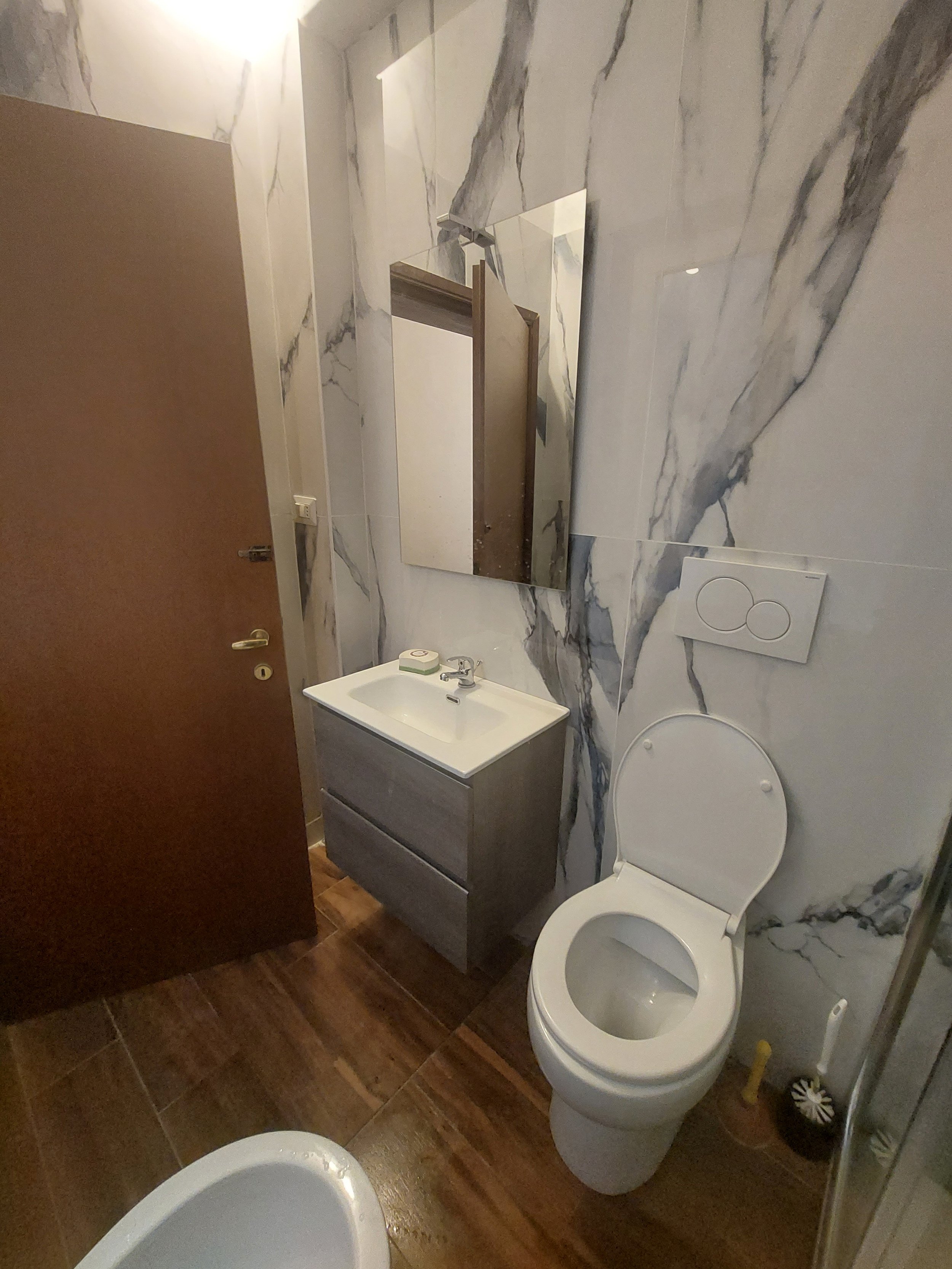 Vista di un bagno con pareti rivestite in marmo bianco con venature grigie, bagno con sanitari, specchio, e mobile con lavabo, in una stanza con pavimento in piastrelle di colore marrone.