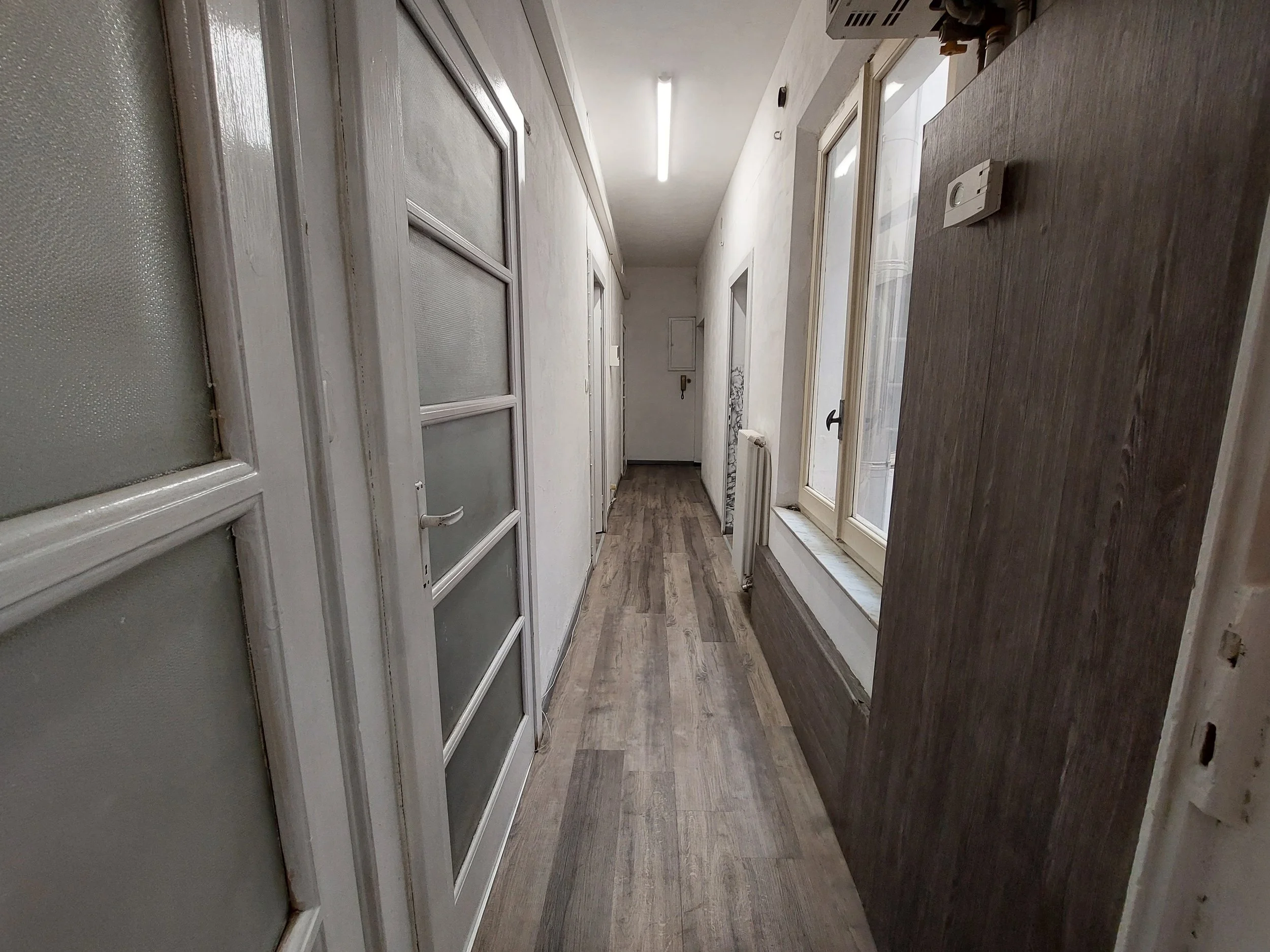Corridoio interno di un edificio con pareti bianche, porte e grandi finestre su un lato e pavimento in legno scuro.