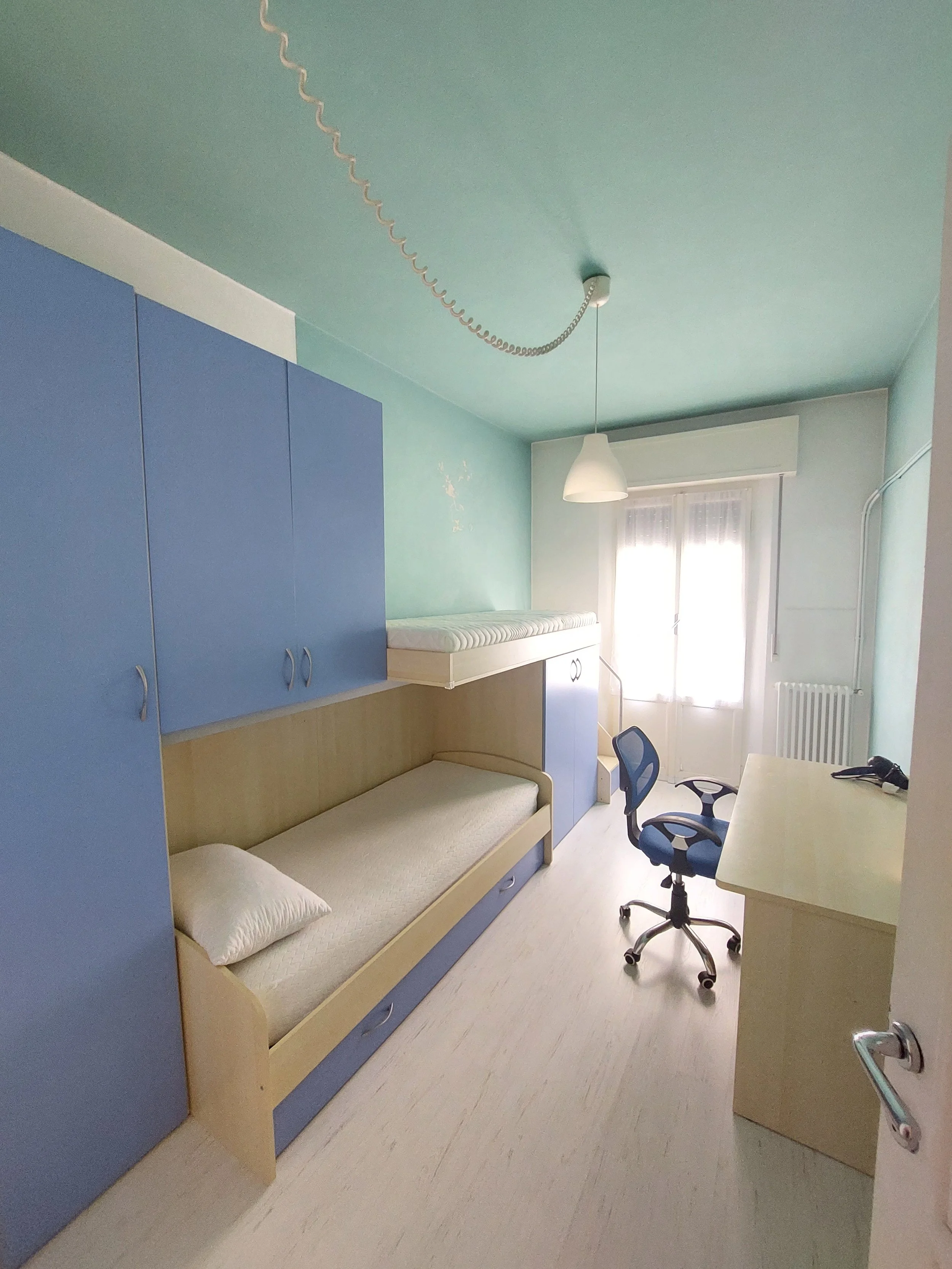Una stanza con un letto a castello, armadi blu, una scrivania e una sedia da ufficio, con molta luce naturale che entra dalle finestre.