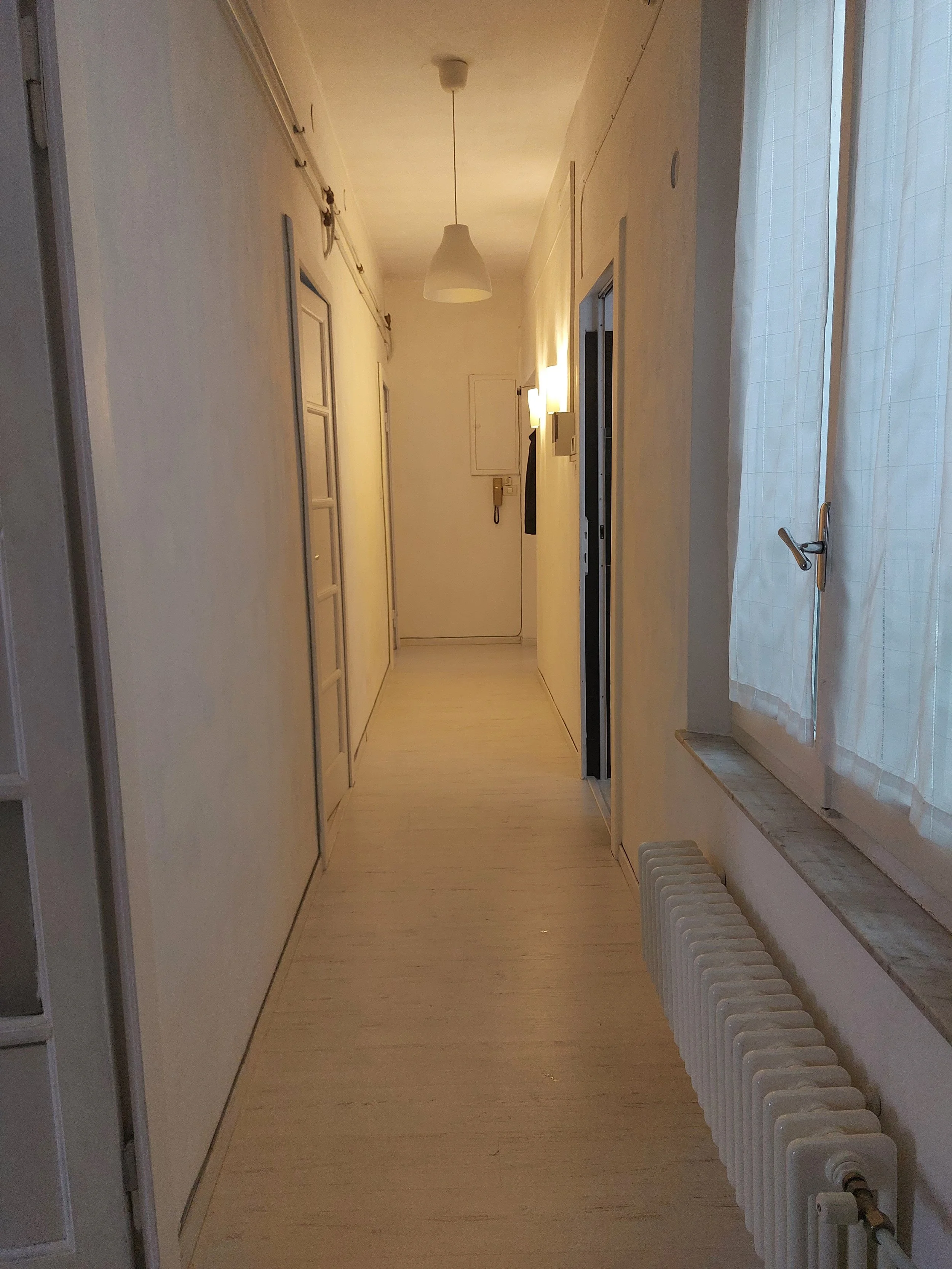 Corridor di casa con pareti bianche, vetri con tende bianche, termosifoni sulla parete destro e alcune luci accese sulle pareti.