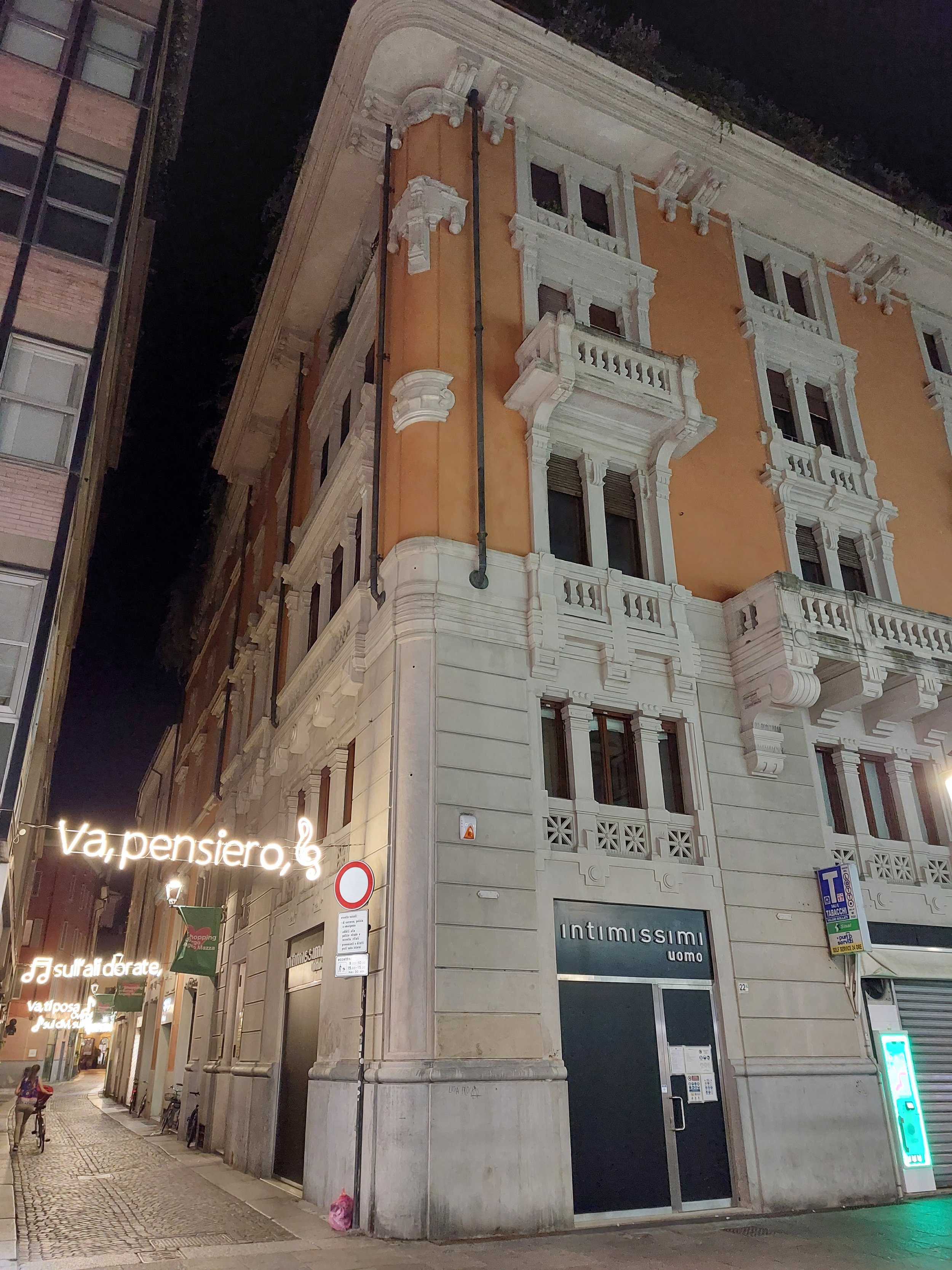 Immagine di un edificio storico con facciata in stile classico, balconi con ringhiere in pietra, edicola luminosa sul marciapiede, strada pedonale e persone che camminano, notturna.