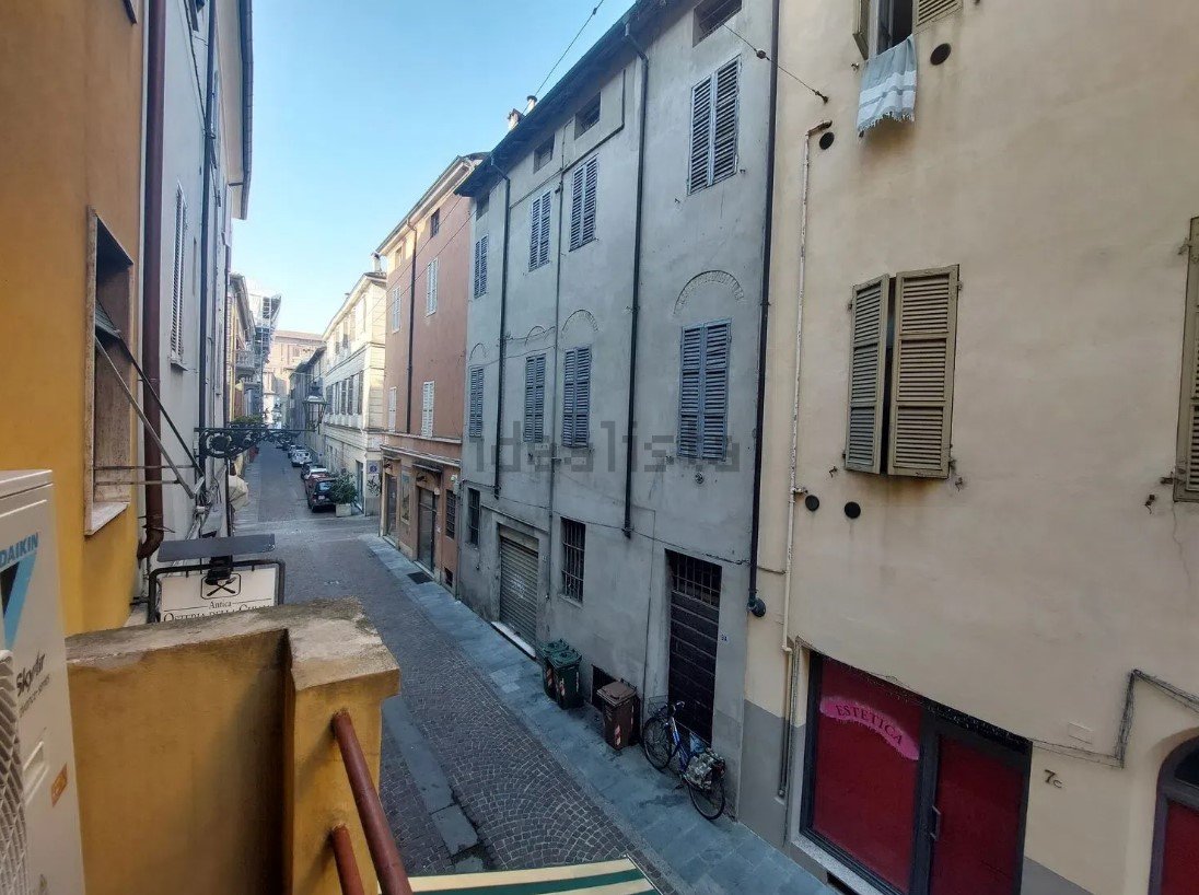 Vista di una strada stretta in un quartiere residenziale con edifici adiacenti di tre o quattro piani, alcune finestre con persiane, biciclette e bidoni della spazzatura lungo il marciapiede, in un giorno soleggiato.